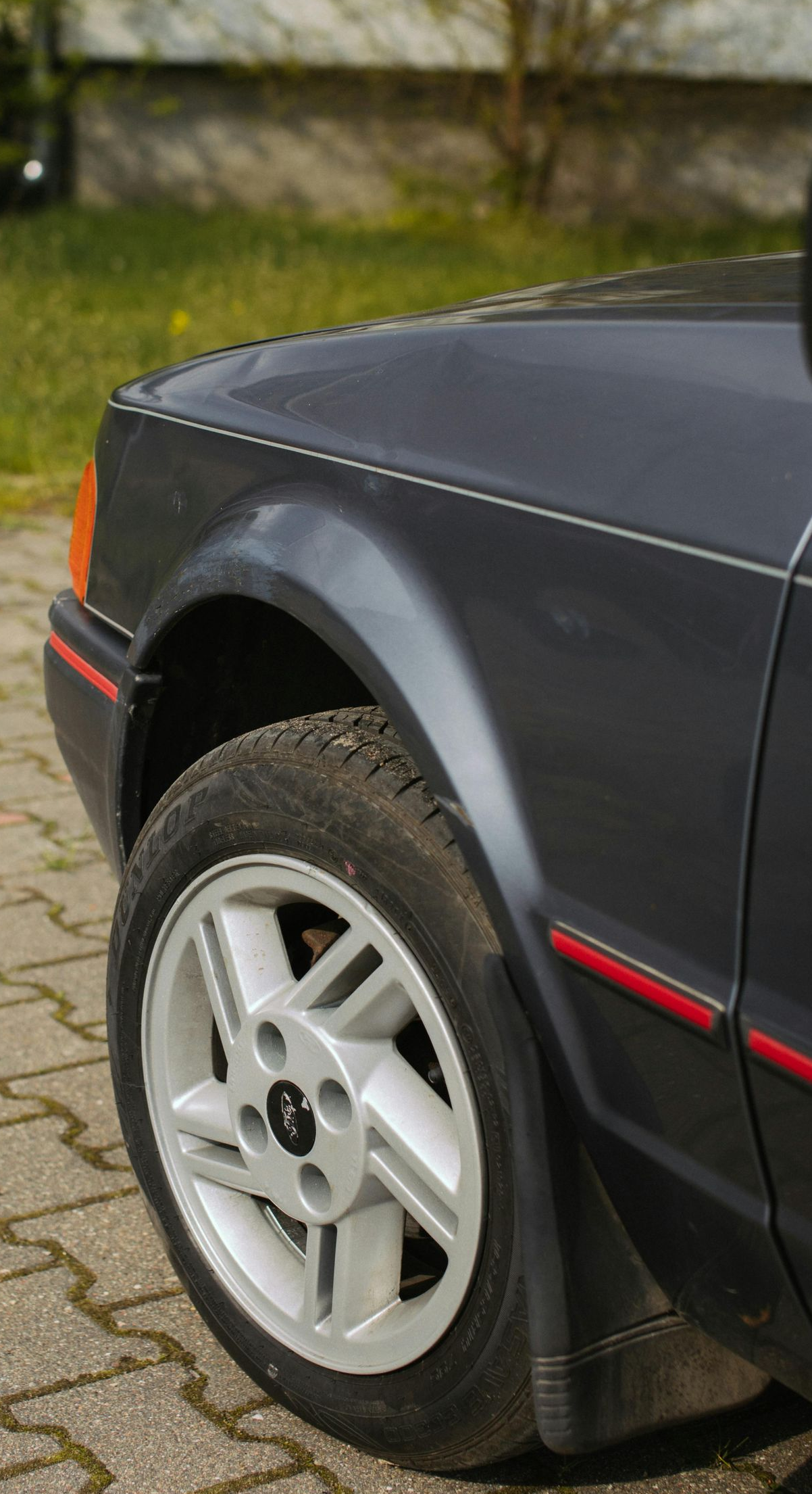 Dark grey Ford Escort front quarter panel, wheel on brick pavement.
