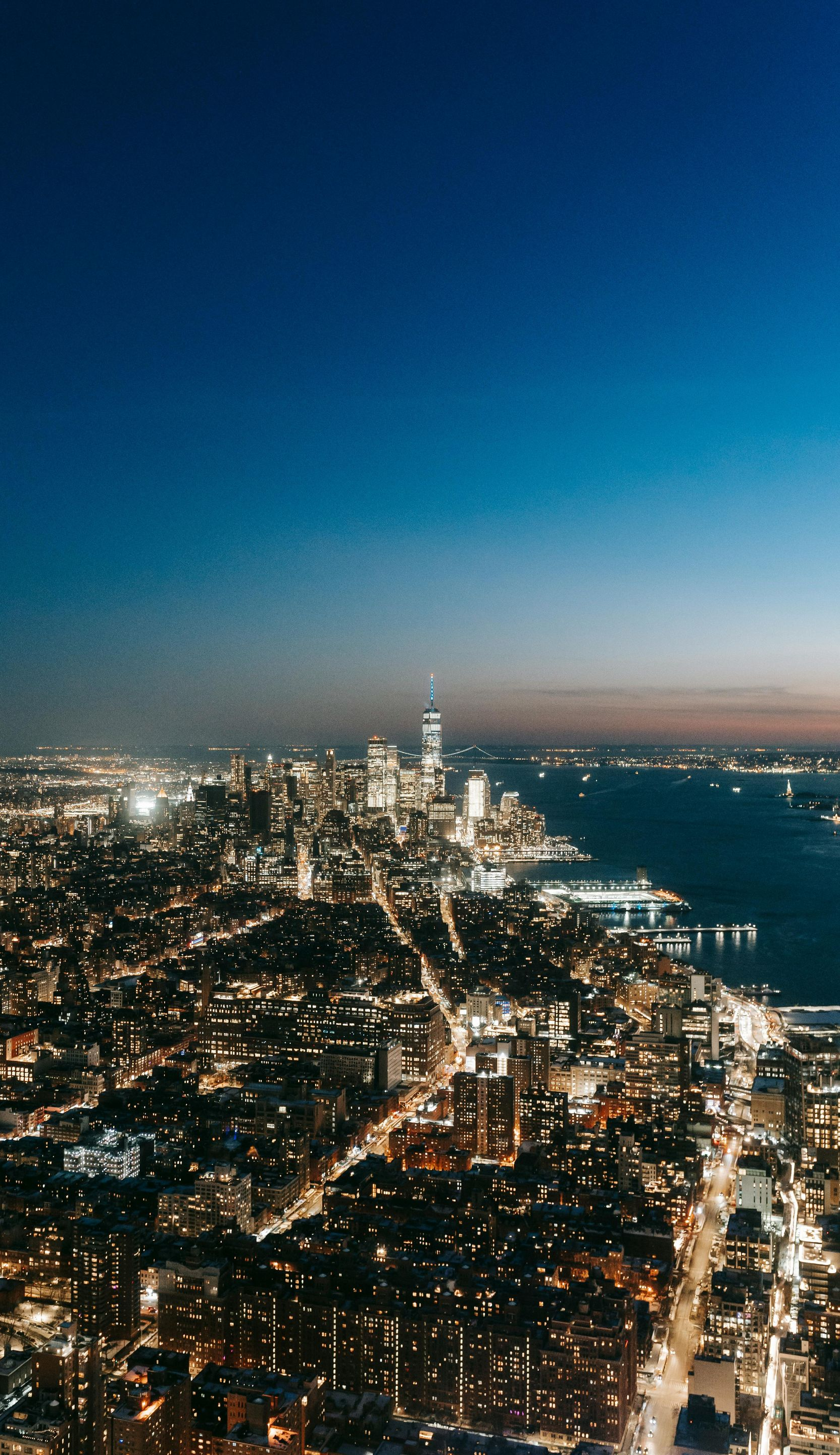 Night view of a cityscape with bright lights, a river, and a dark blue sky.
