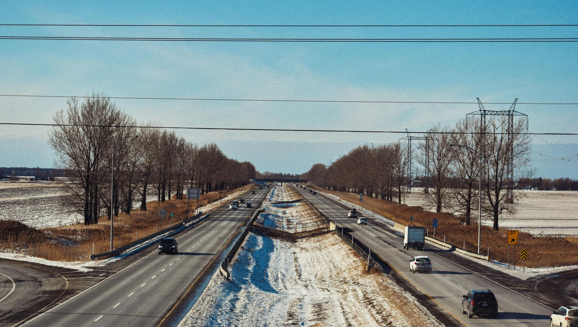 Highway with cars and trucks, flanked by trees and snow-covered ground, under a blue sky.