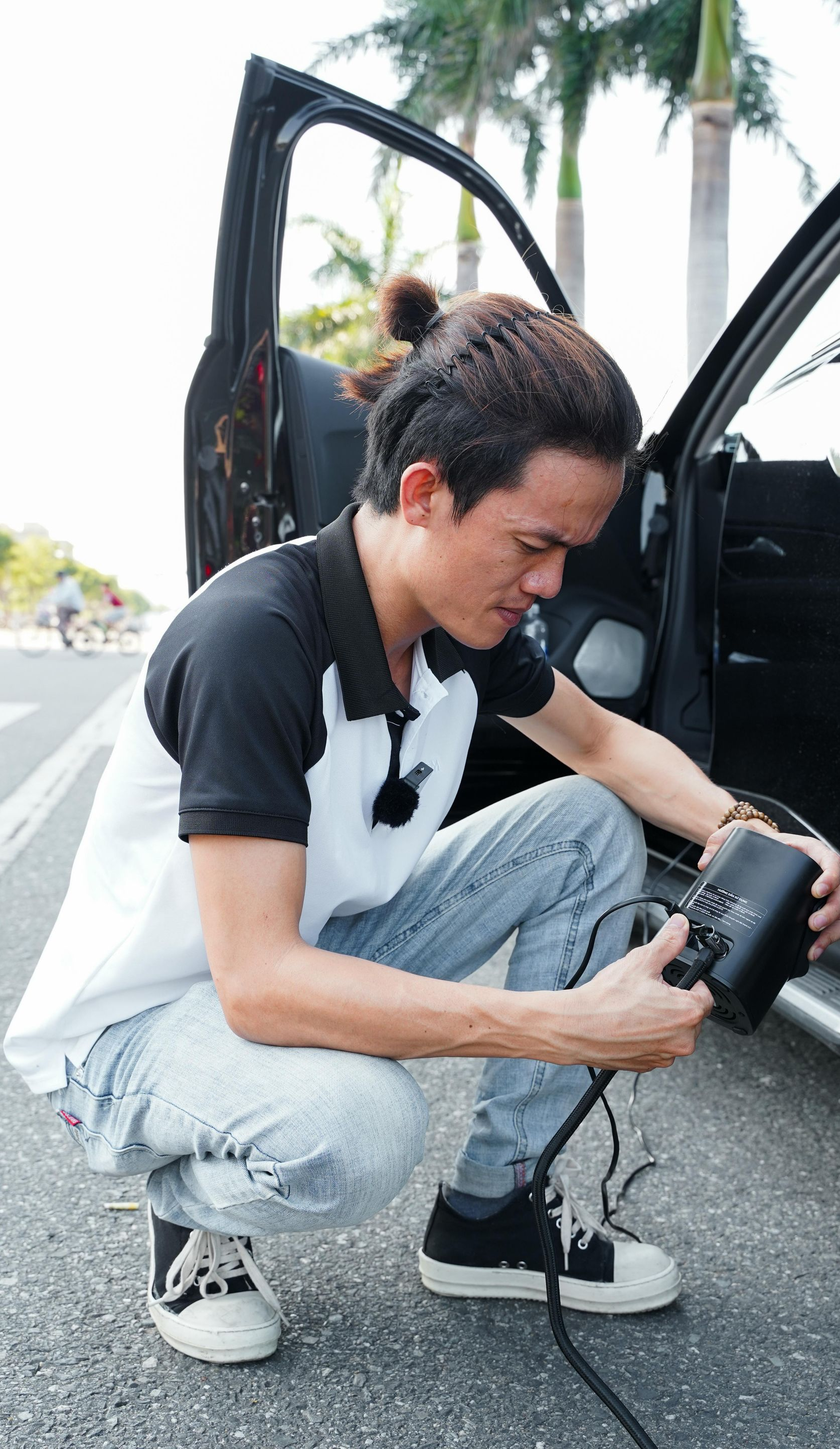 Man crouches next to a car, connecting a black cable. Open car door, outdoor setting.