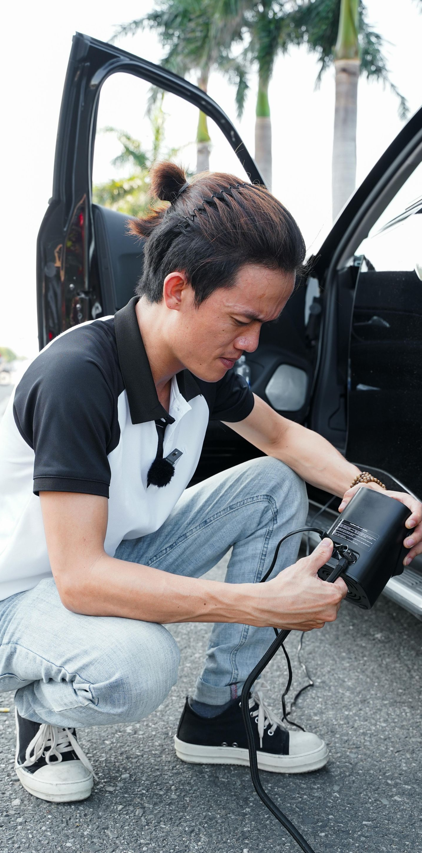 Man crouches next to a car, connecting a black cable. Open car door, outdoor setting.