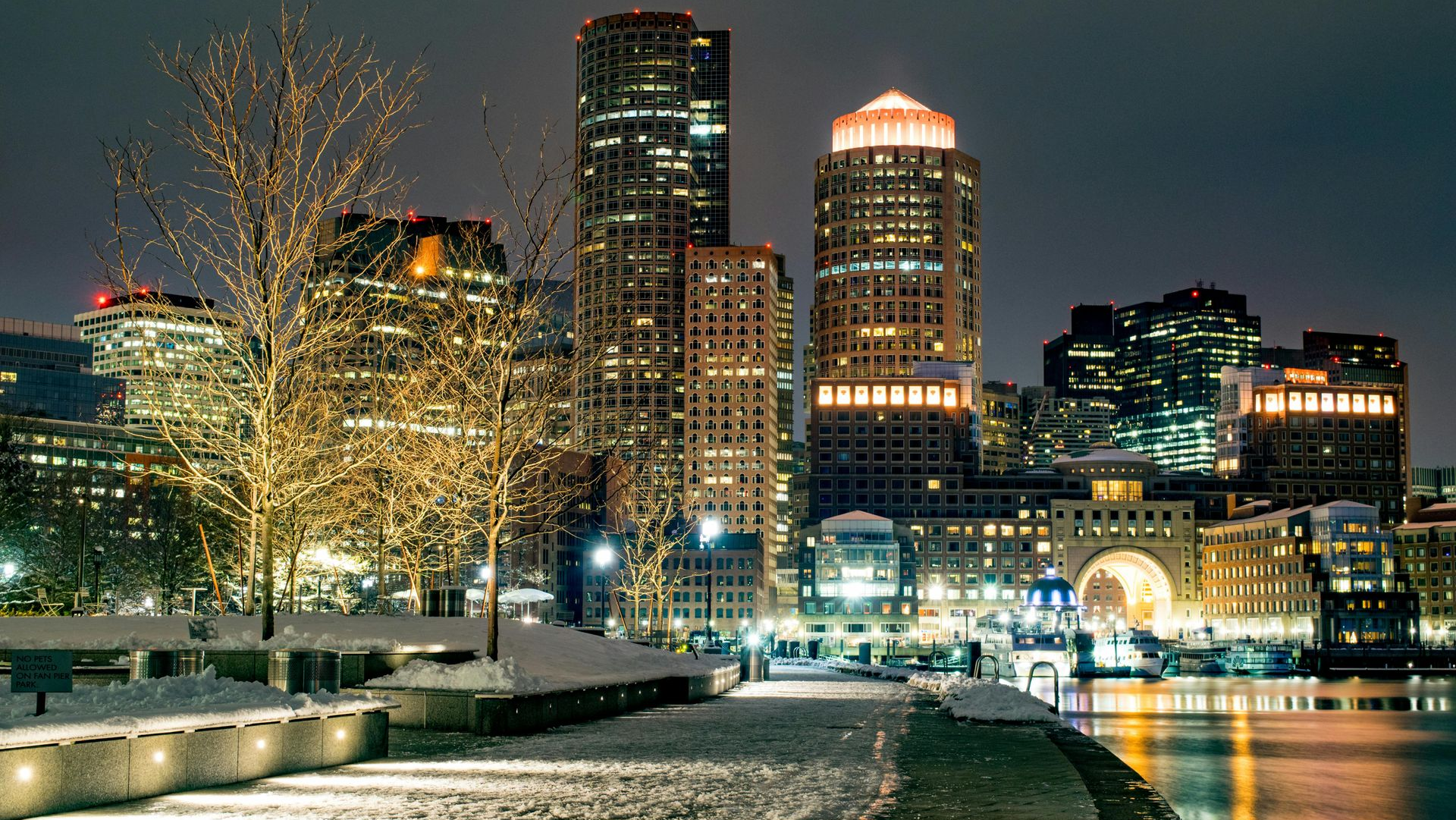 Night view of Boston skyline with lit buildings and snow-covered foreground along the water.
