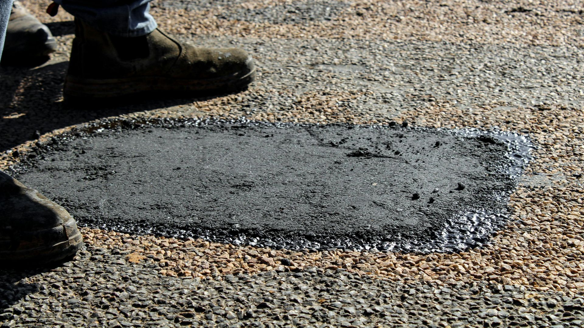 A person’s feet stand on a gravel surface next to a freshly filled asphalt patch on a road.