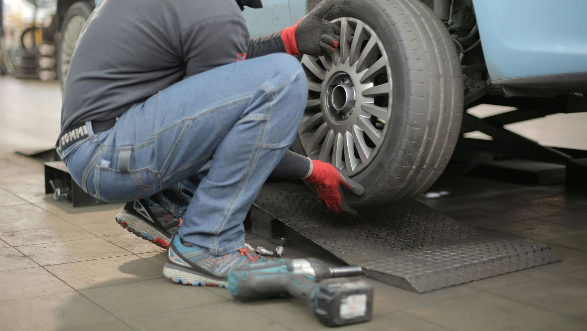 Mechanic changing a tire on a blue car in a garage, using a power tool and wearing red gloves.