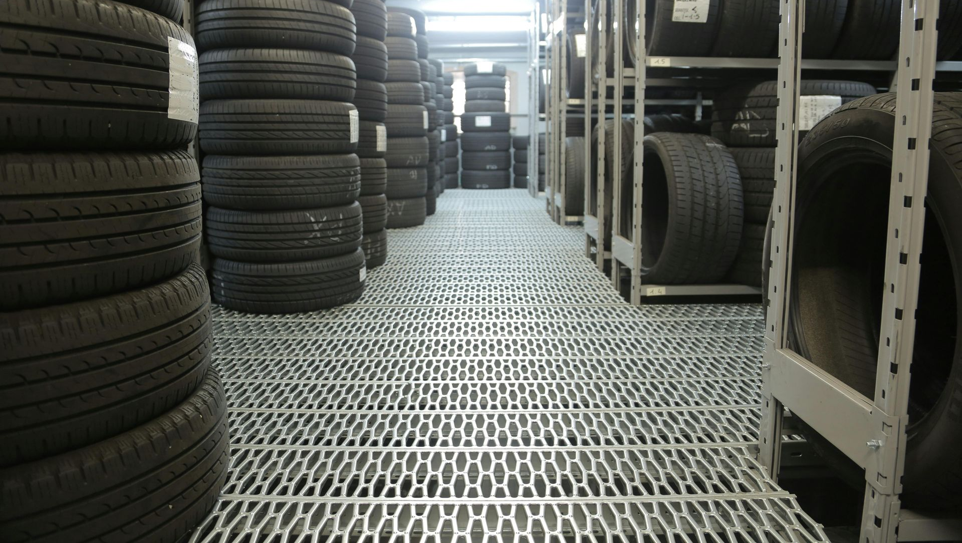 A perspective view of a warehouse storage aisle filled with stacks of black tires on metal flooring and shelves.