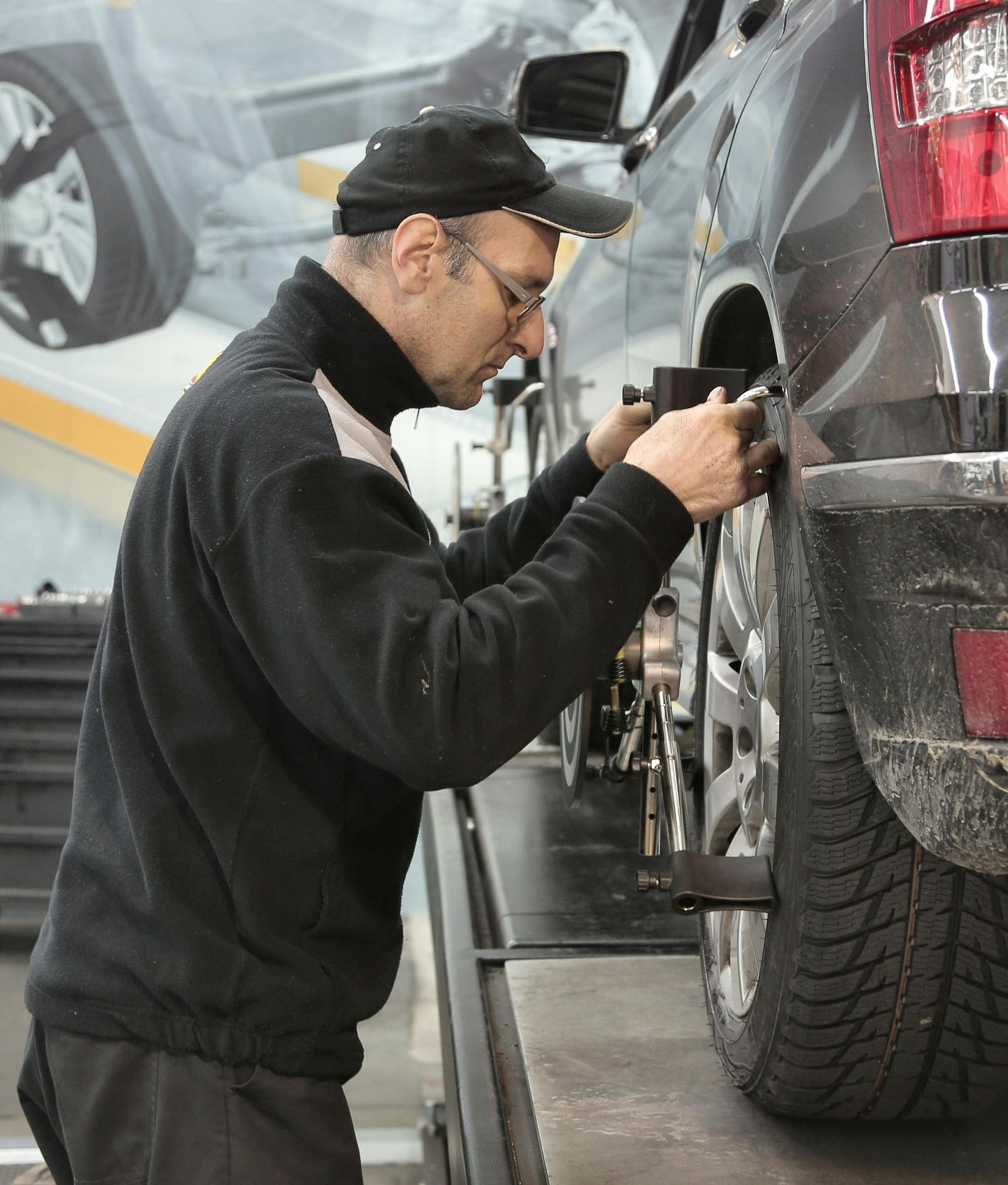 Mechanic adjusts car wheel alignment. He wears a black jacket and hat, working near a tire.