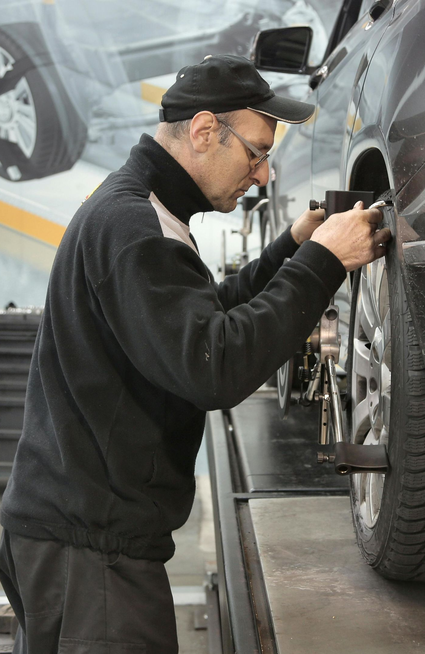 Mechanic aligning car wheel on a lift, wearing a cap and glasses.