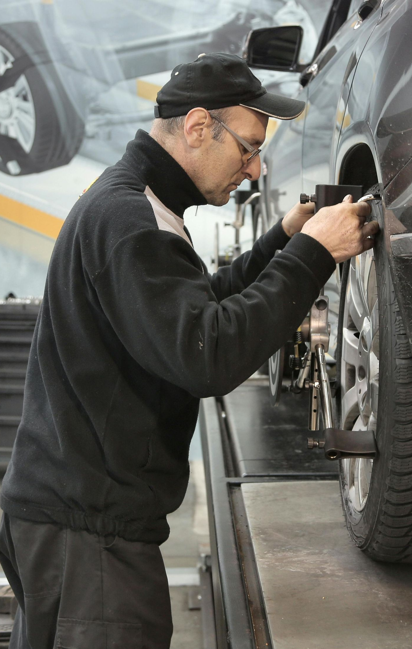 Mechanic aligning car wheel on a lift, wearing a cap and glasses.