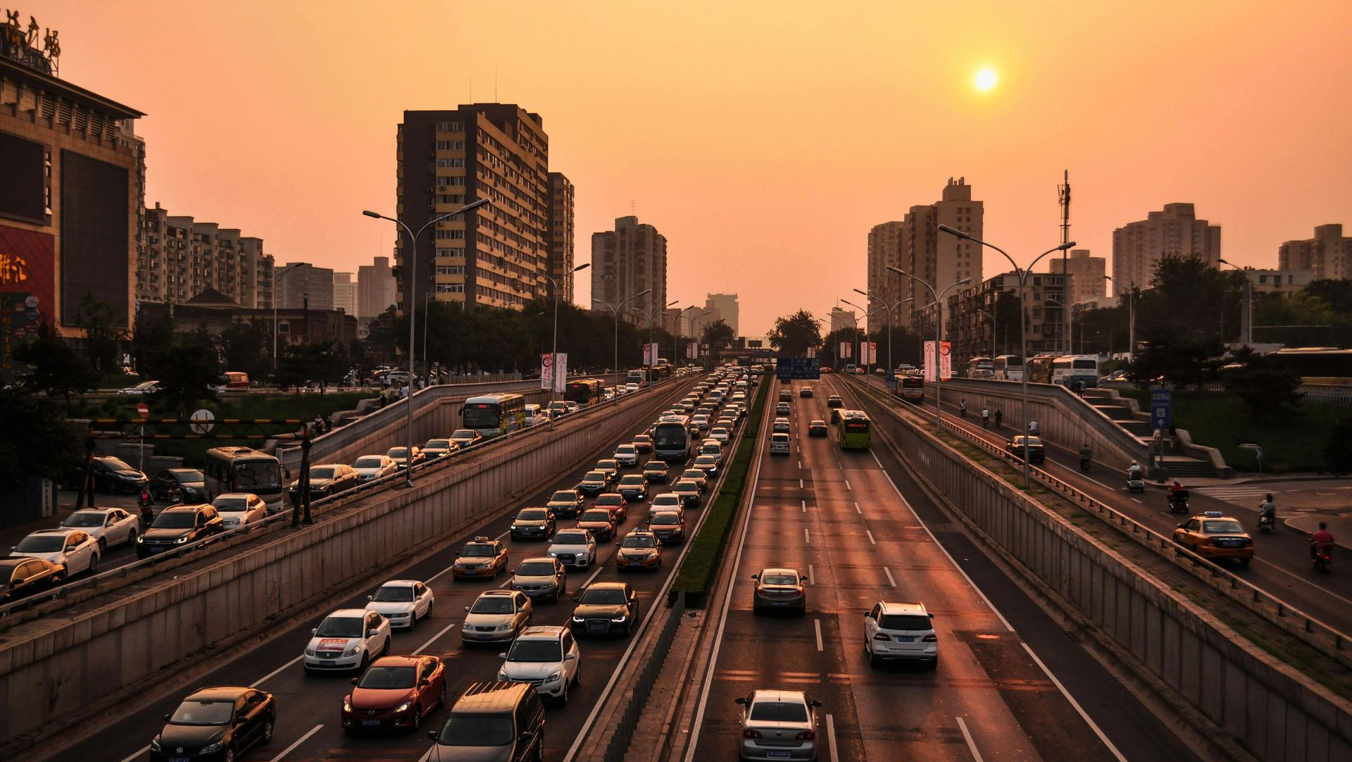 Traffic jam on a multi-lane highway in a city at sunset; tall buildings in background.
