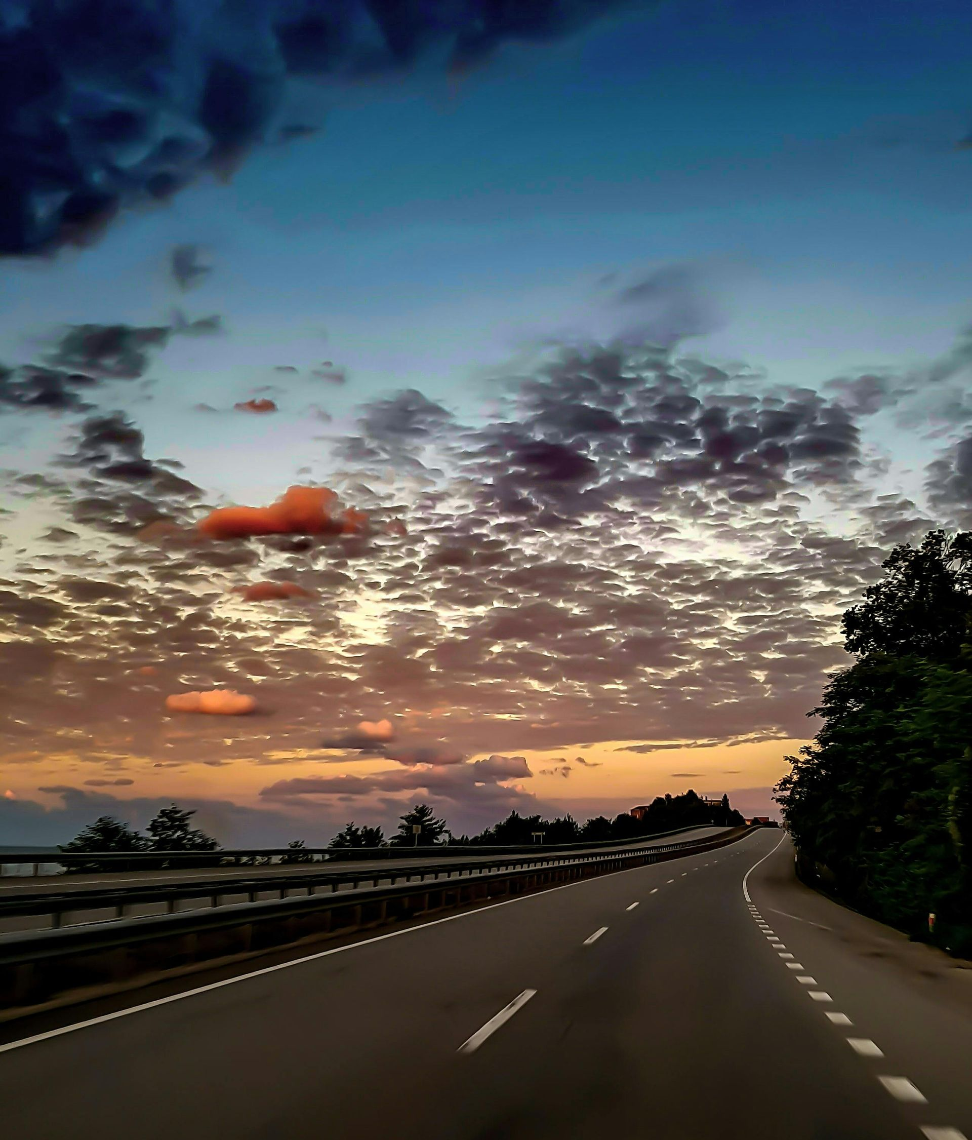 Road curving into the distance at sunset, under a sky of clouds and orange, blue, and purple hues.