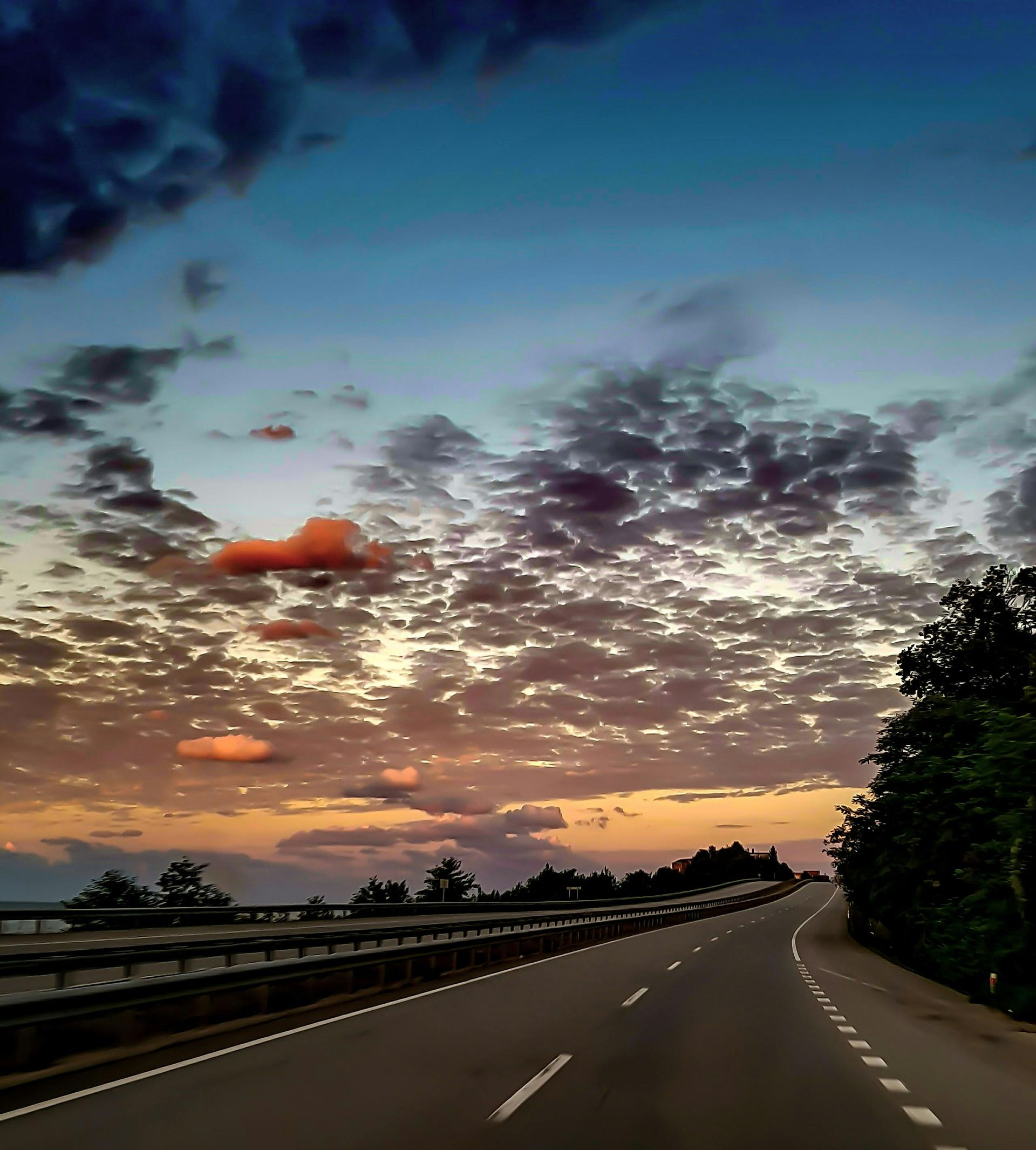 Road curving into the distance at sunset, under a sky of clouds and orange, blue, and purple hues.