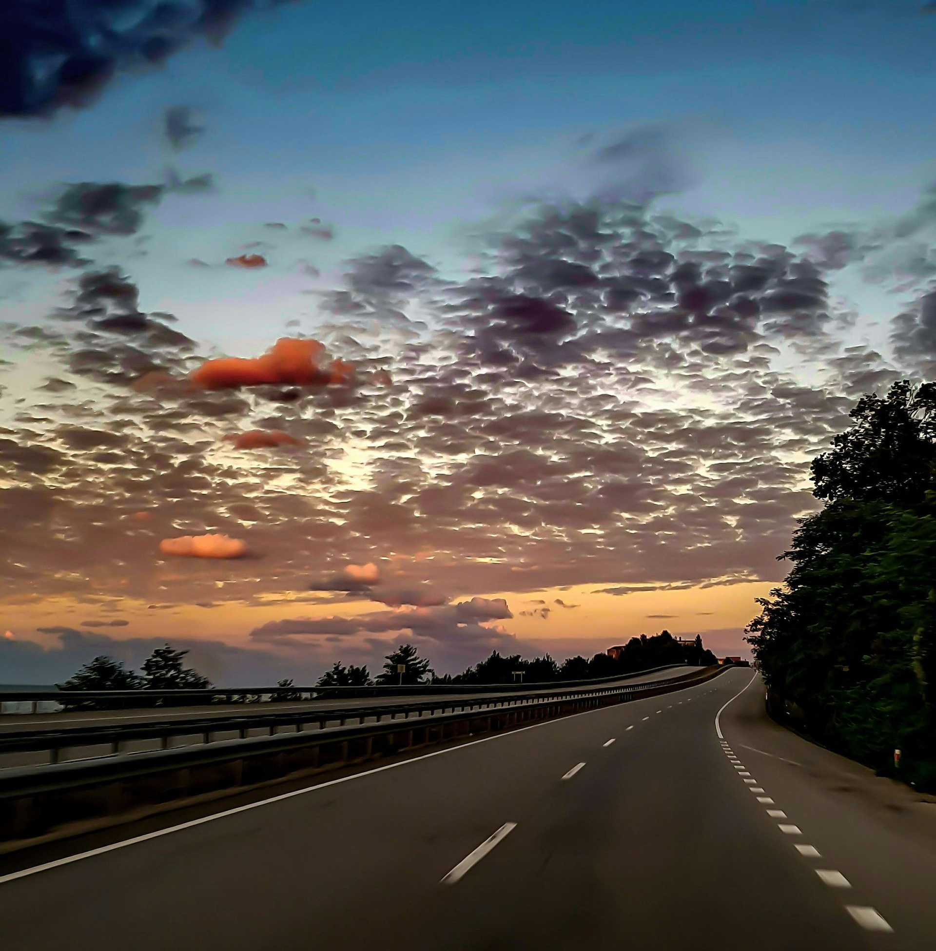 Road curving into the distance at sunset, under a sky of clouds and orange, blue, and purple hues.