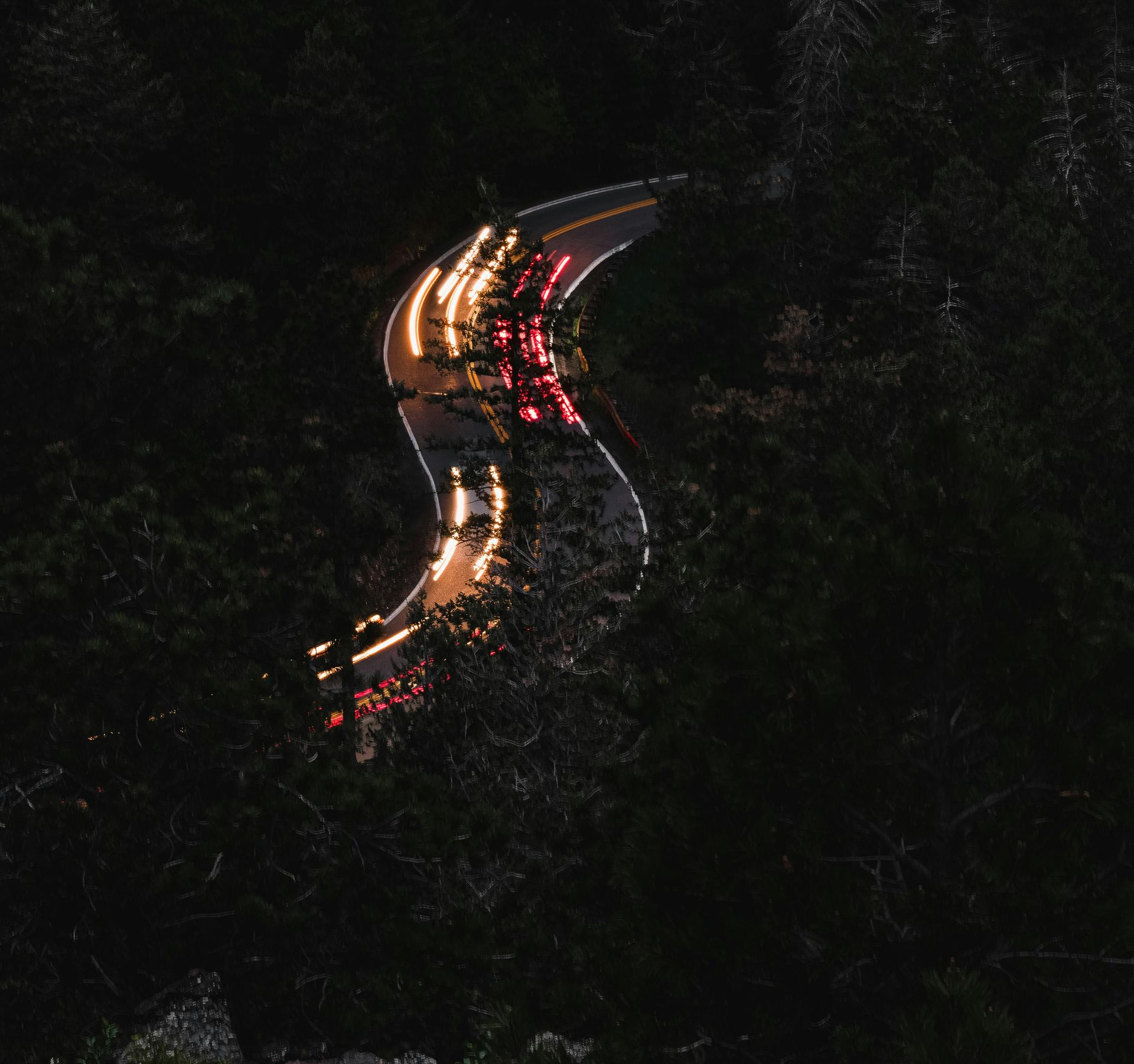 Nighttime aerial view of a winding road with blurred car lights snaking through a dark forest.