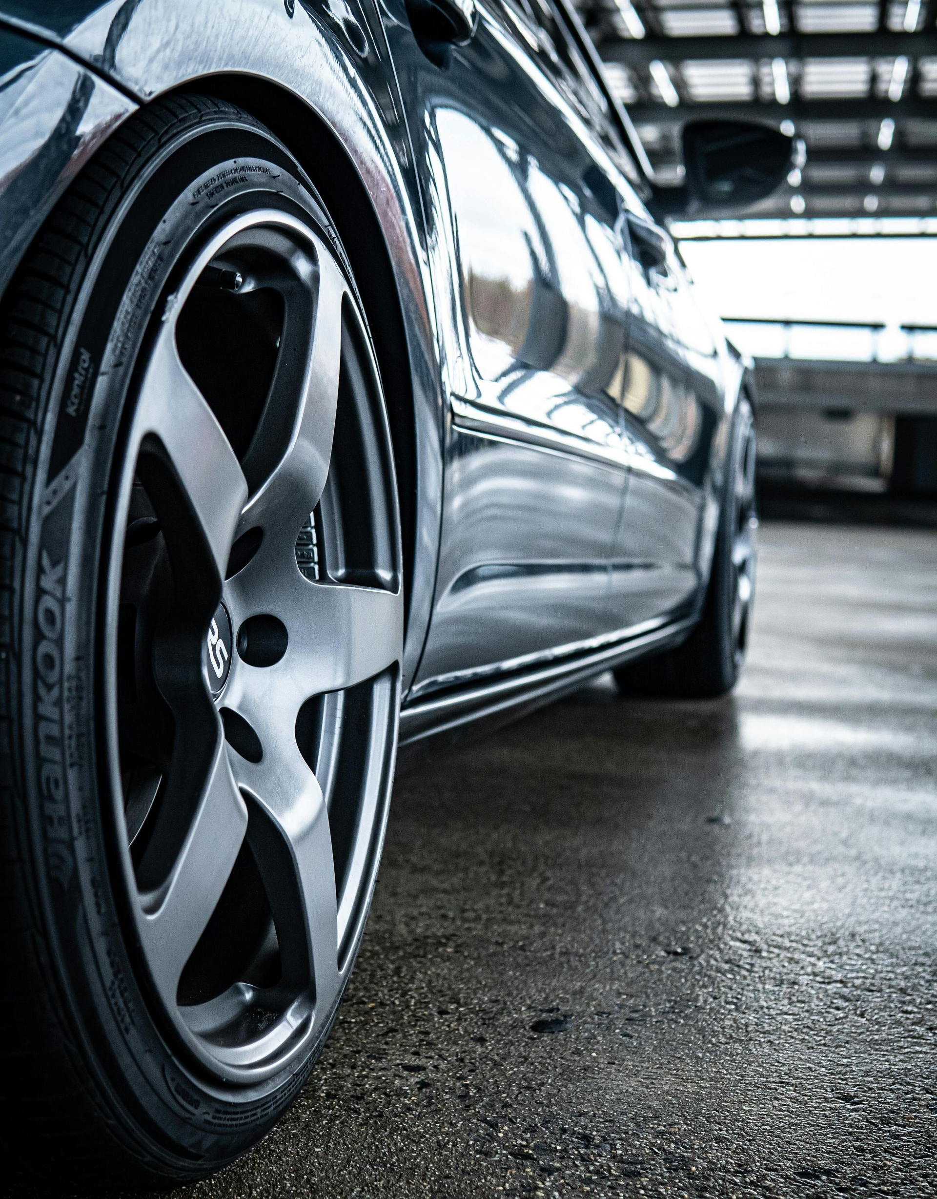 A low-angle, close-up shot of a dark car wheel with a metallic rim parked on a wet, grey concrete surface.