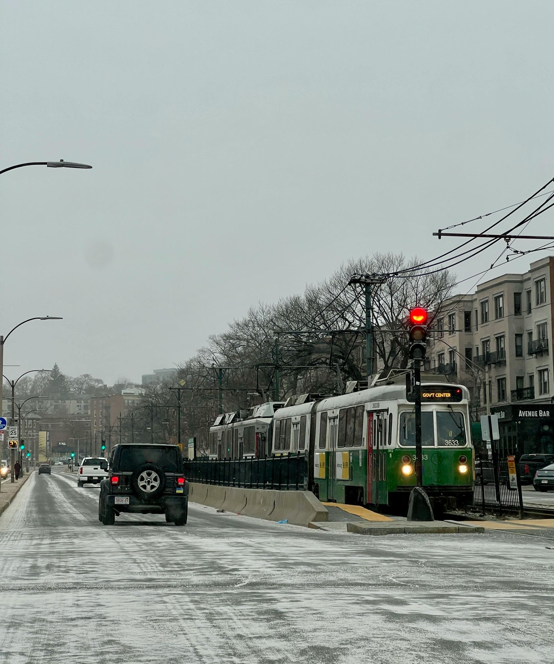 Streetcar stopped at a station on a snow-covered street; cars and buildings in the background.
