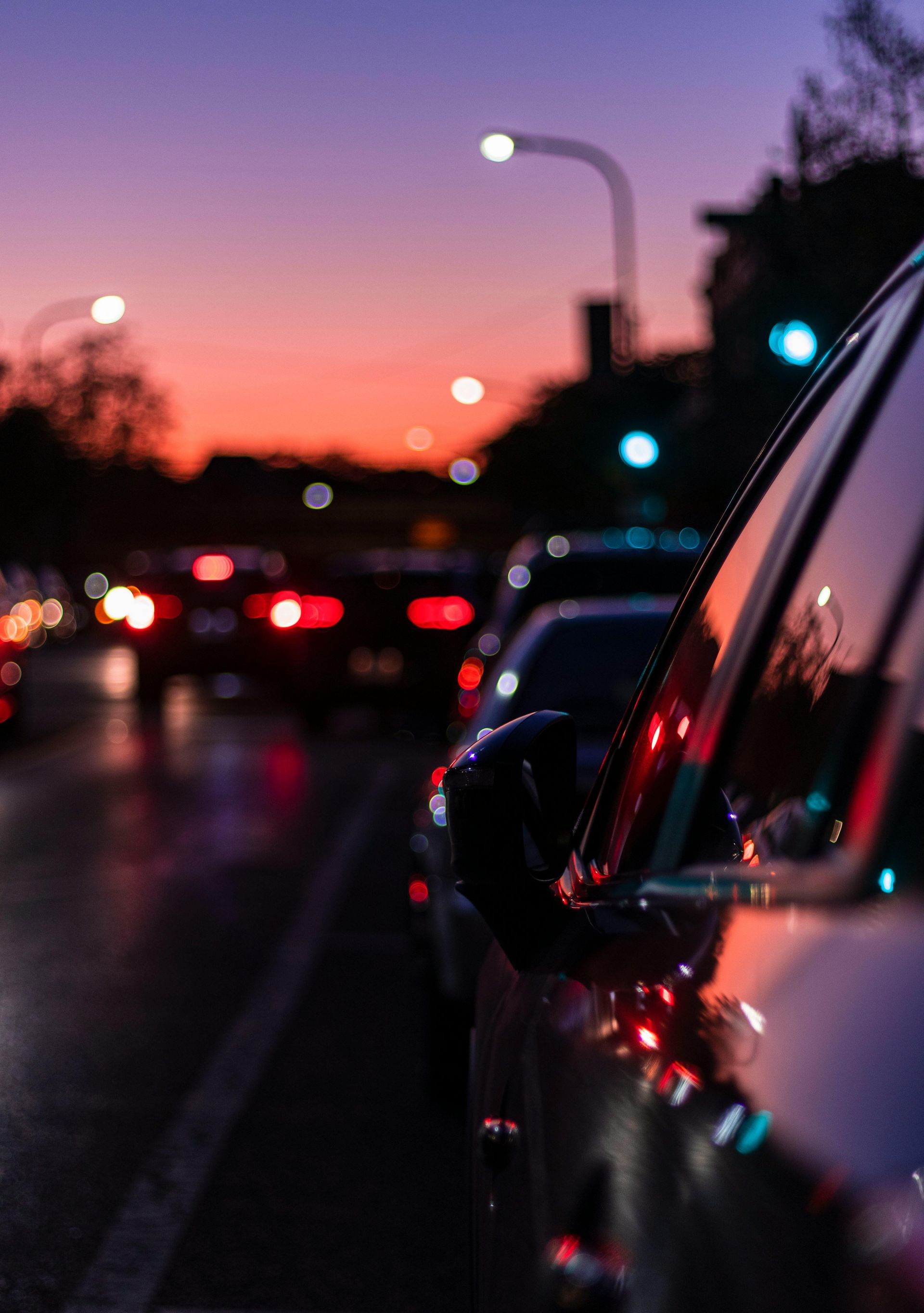 Cars in traffic at dusk, lit by streetlights against a colorful sunset sky.