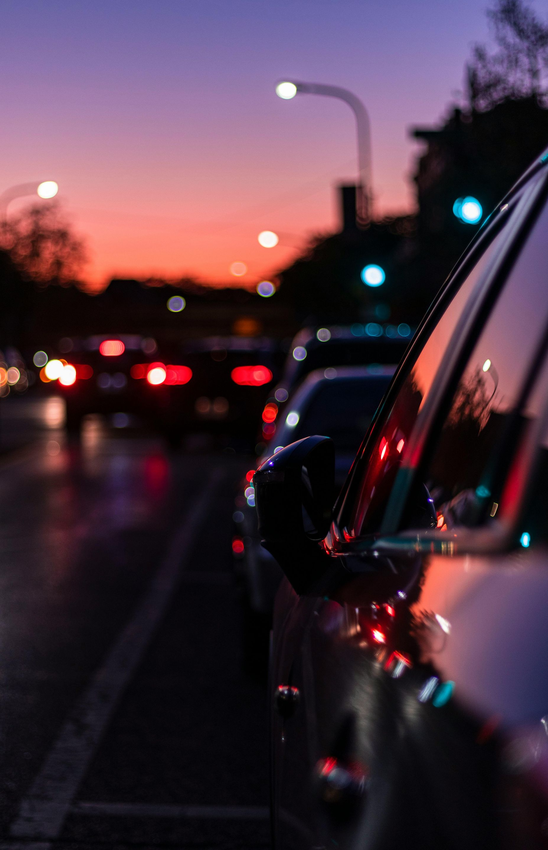 Cars in traffic at dusk, lit by streetlights against a colorful sunset sky.