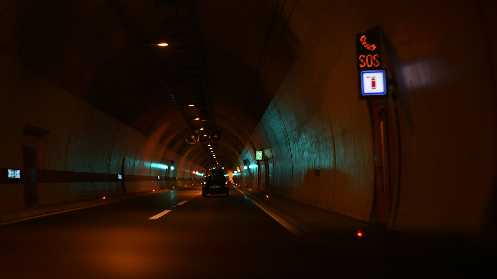 Car driving through a dark tunnel with emergency phone sign lit on the right.