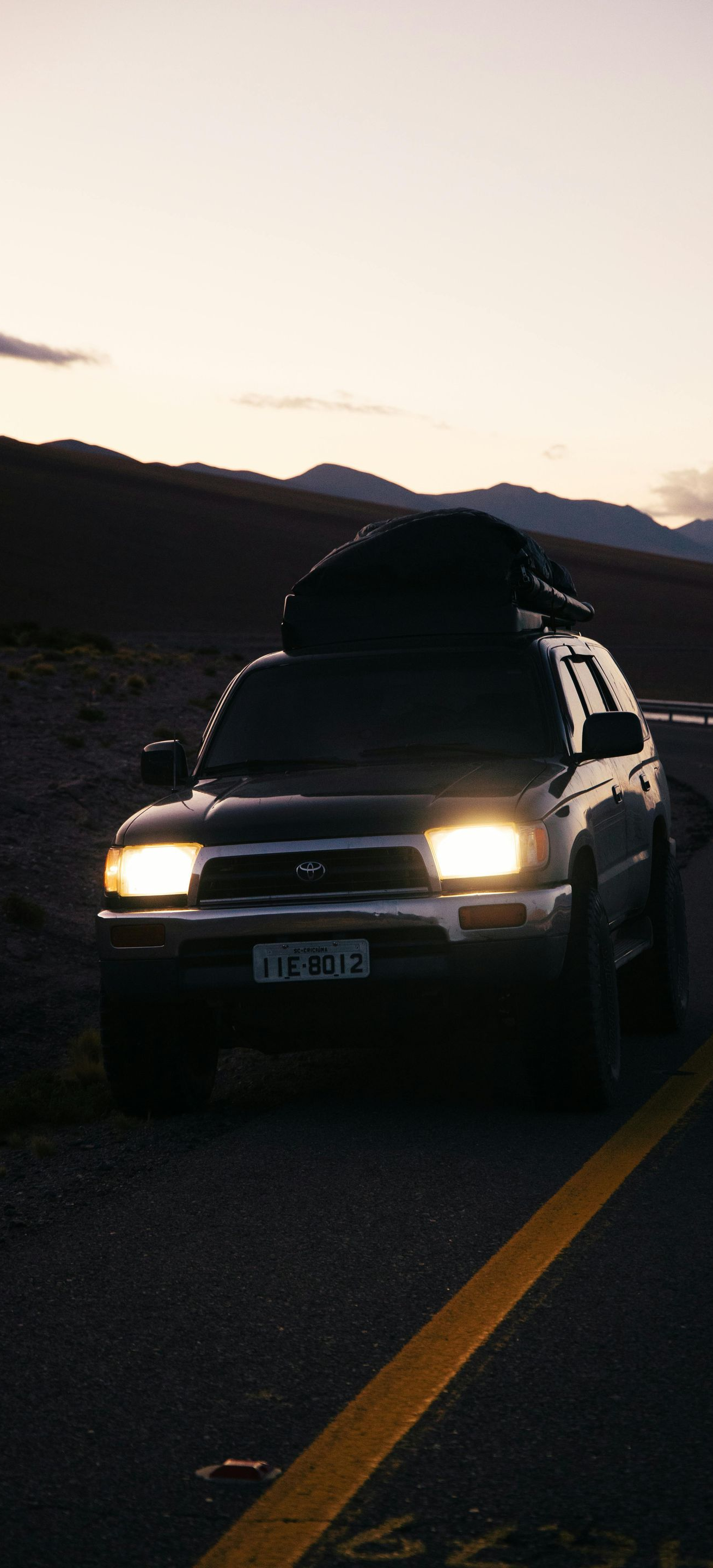 Dark-colored SUV with cargo on its roof parked on a winding road at dusk; mountains in the background.