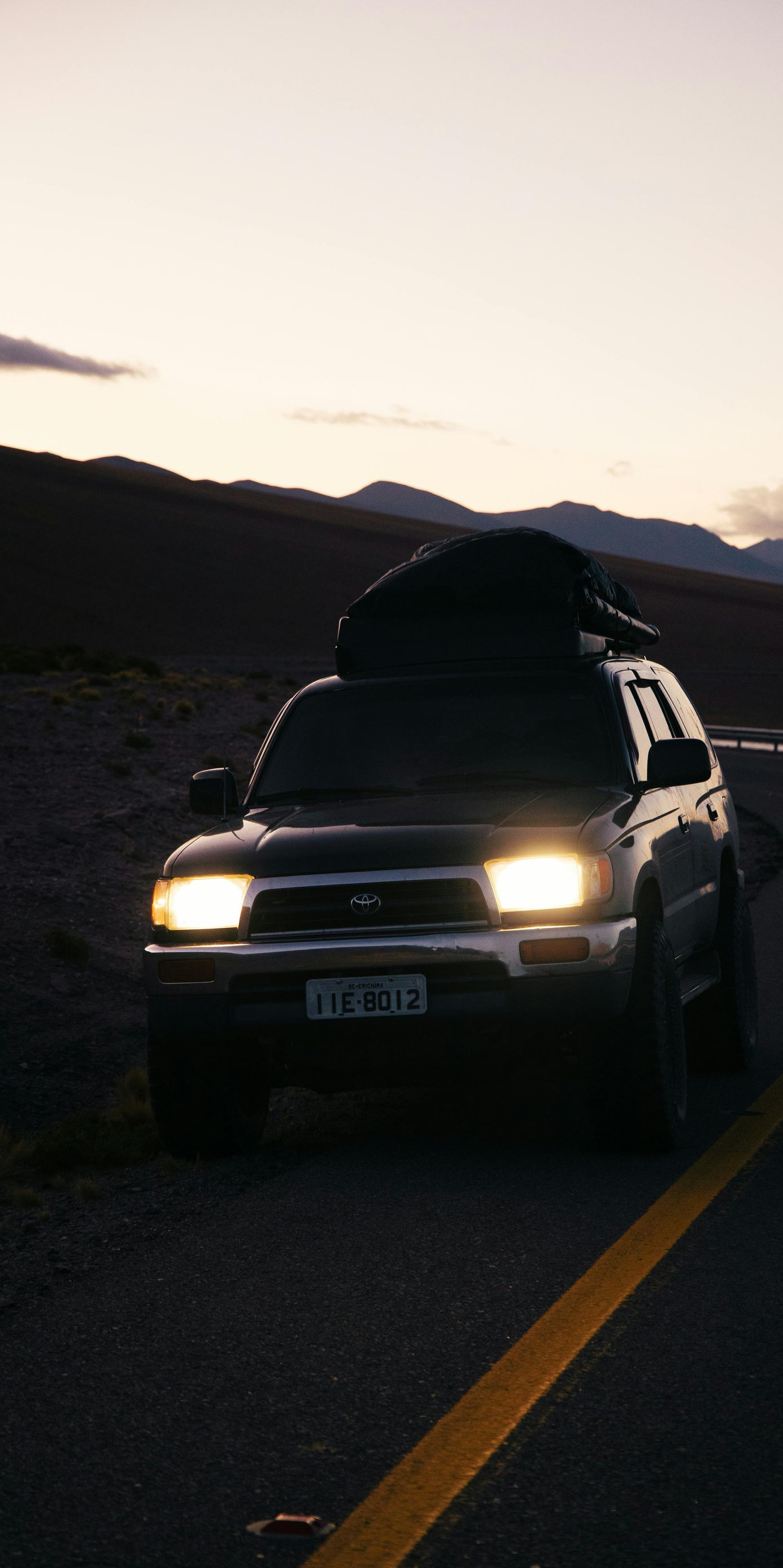 Dark-colored SUV with cargo on its roof parked on a winding road at dusk; mountains in the background.