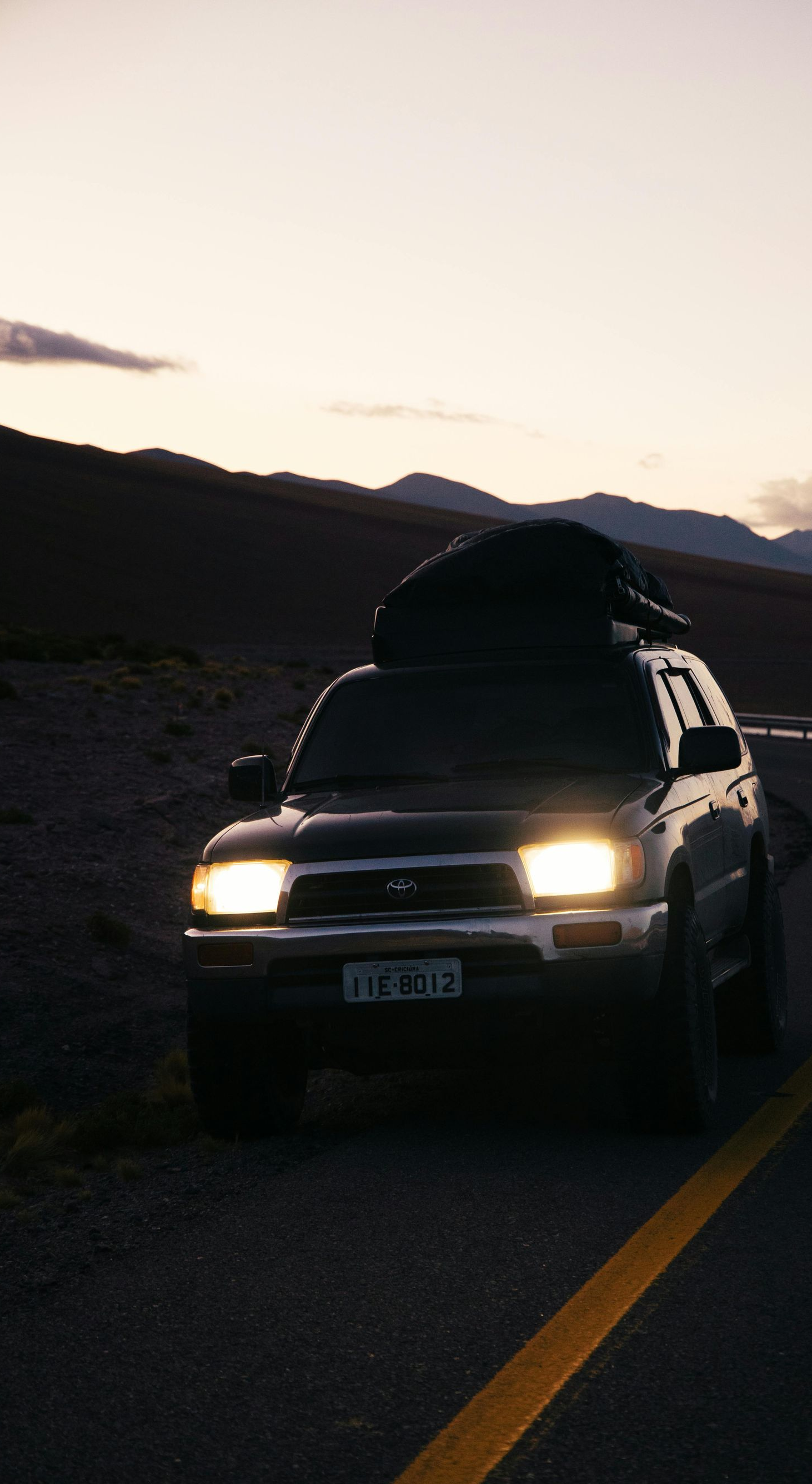 Dark-colored SUV with cargo on its roof parked on a winding road at dusk; mountains in the background.