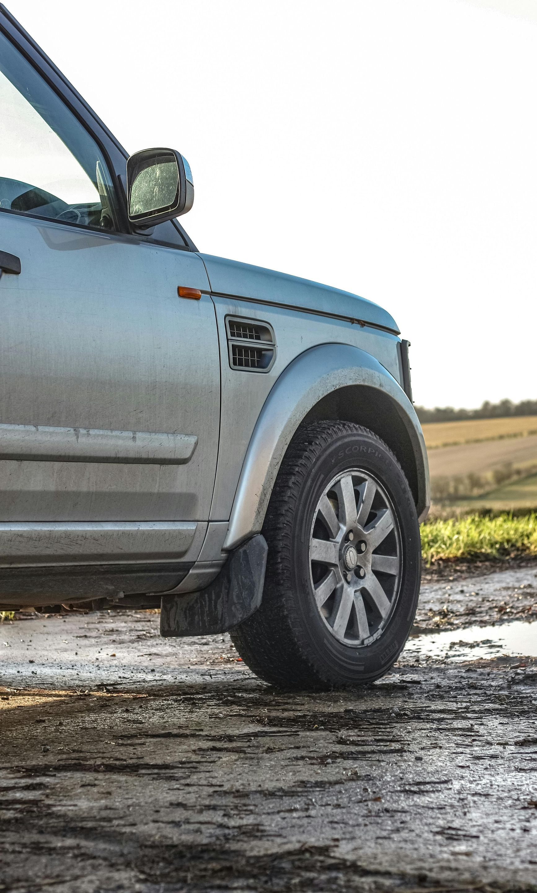 Silver Land Rover SUV on a muddy track, near a field. Partially visible, sunlit.