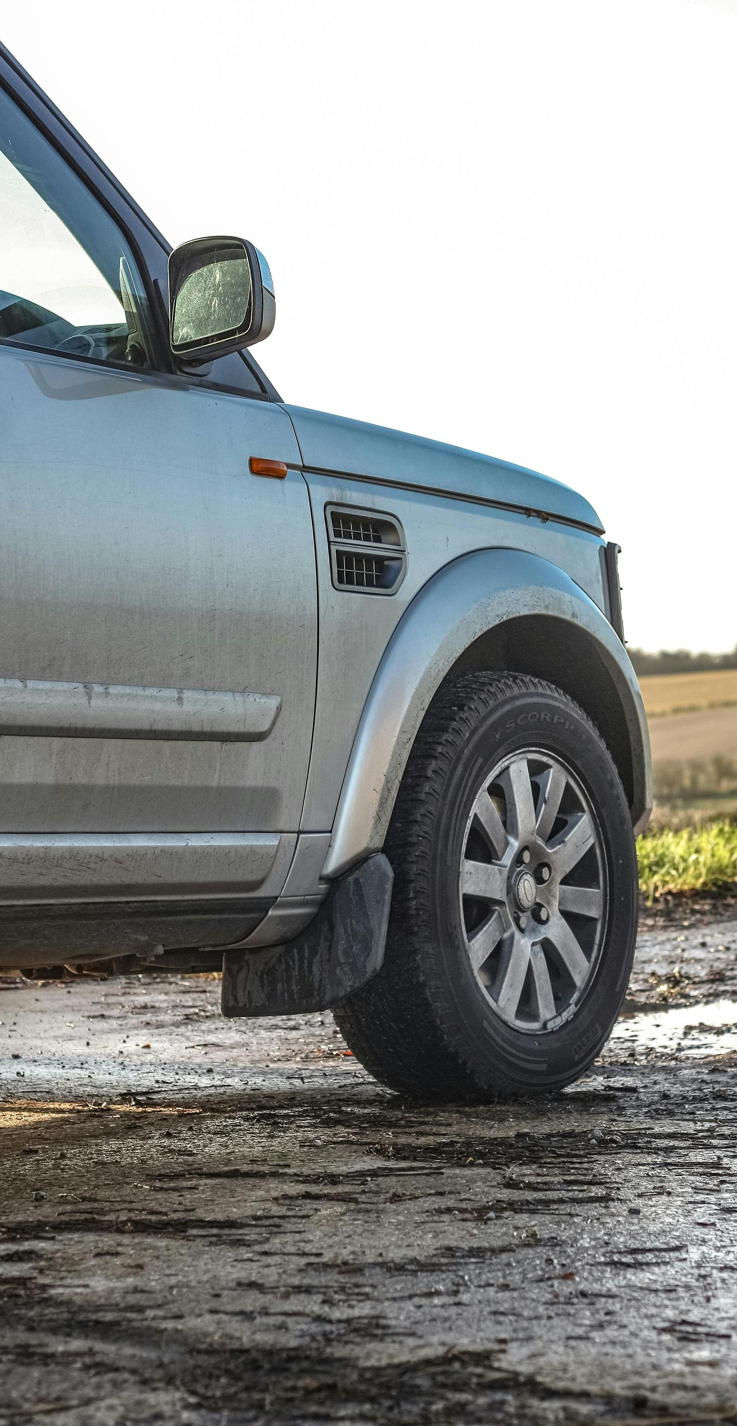 Silver Land Rover SUV on a muddy track, near a field. Partially visible, sunlit.