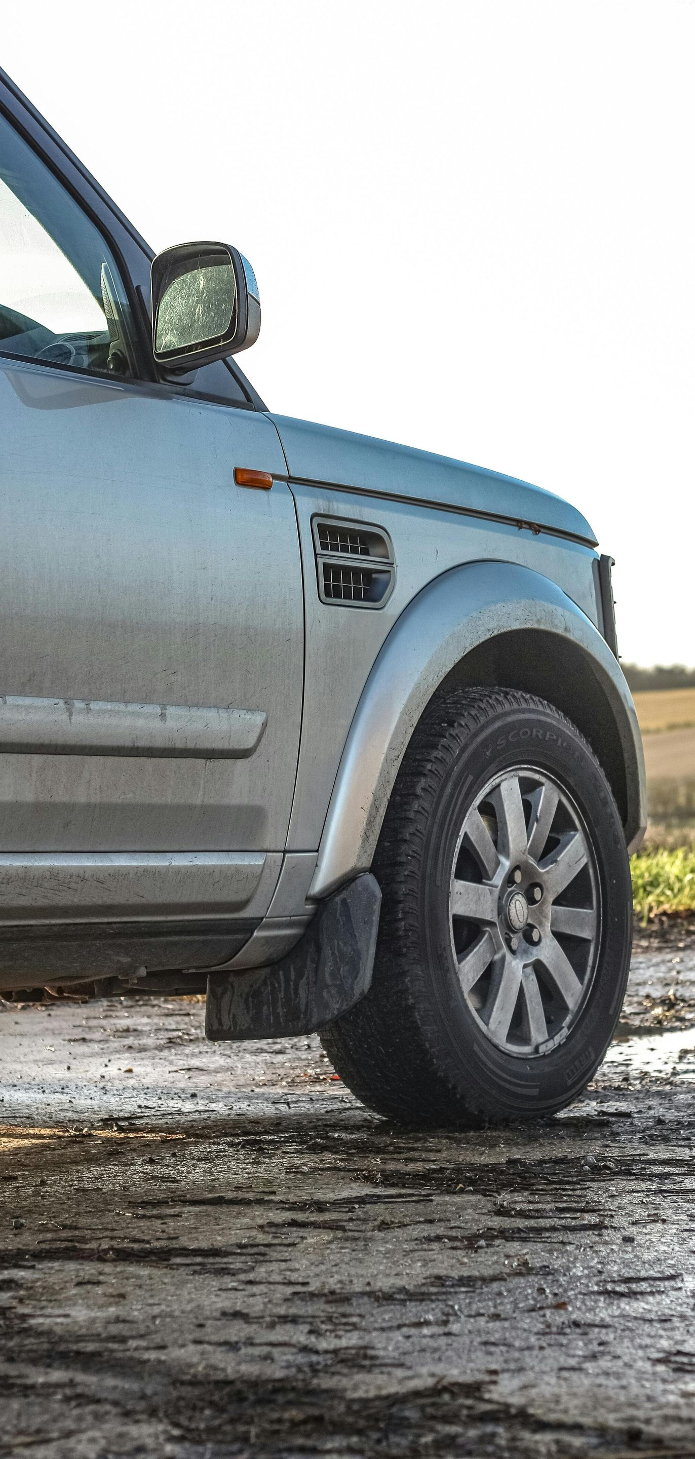 Silver Land Rover SUV on a muddy track, near a field. Partially visible, sunlit.