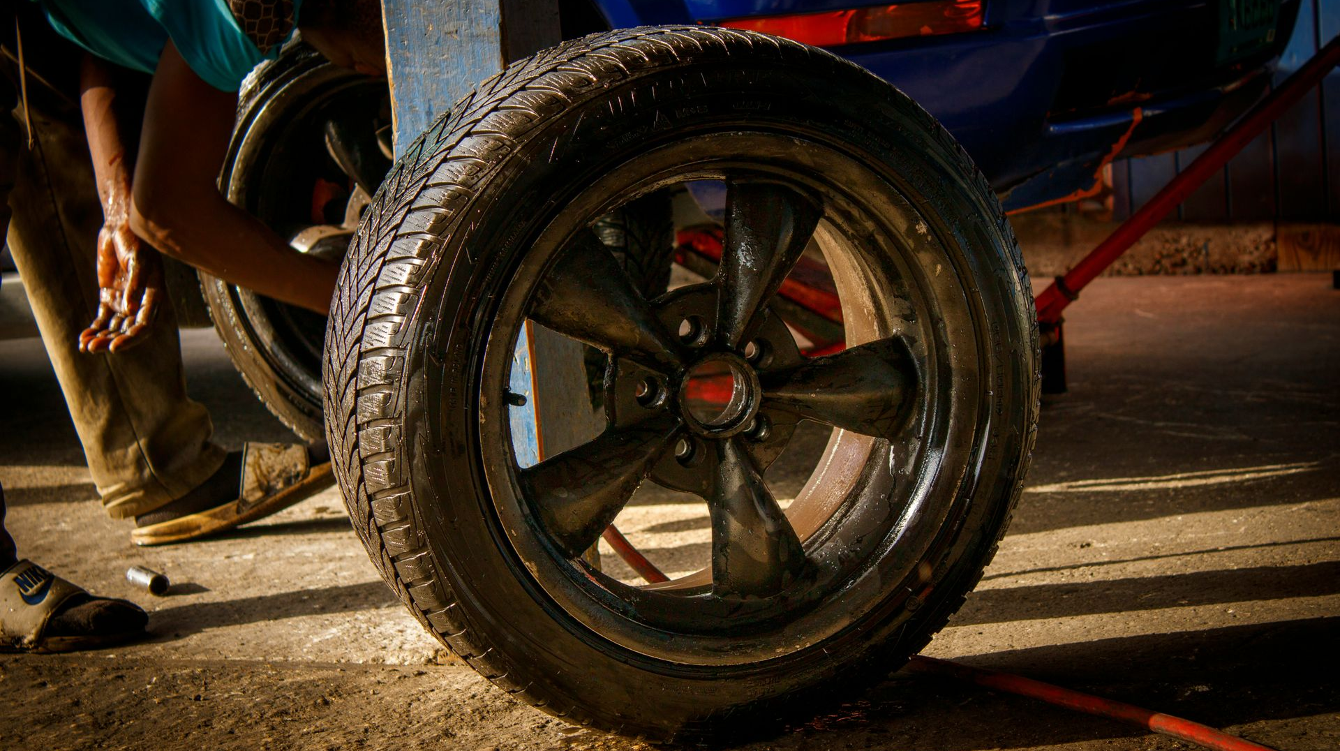 A mechanic works on a tire and five-spoke wheel assembly on a car raised by a jack in a garage.