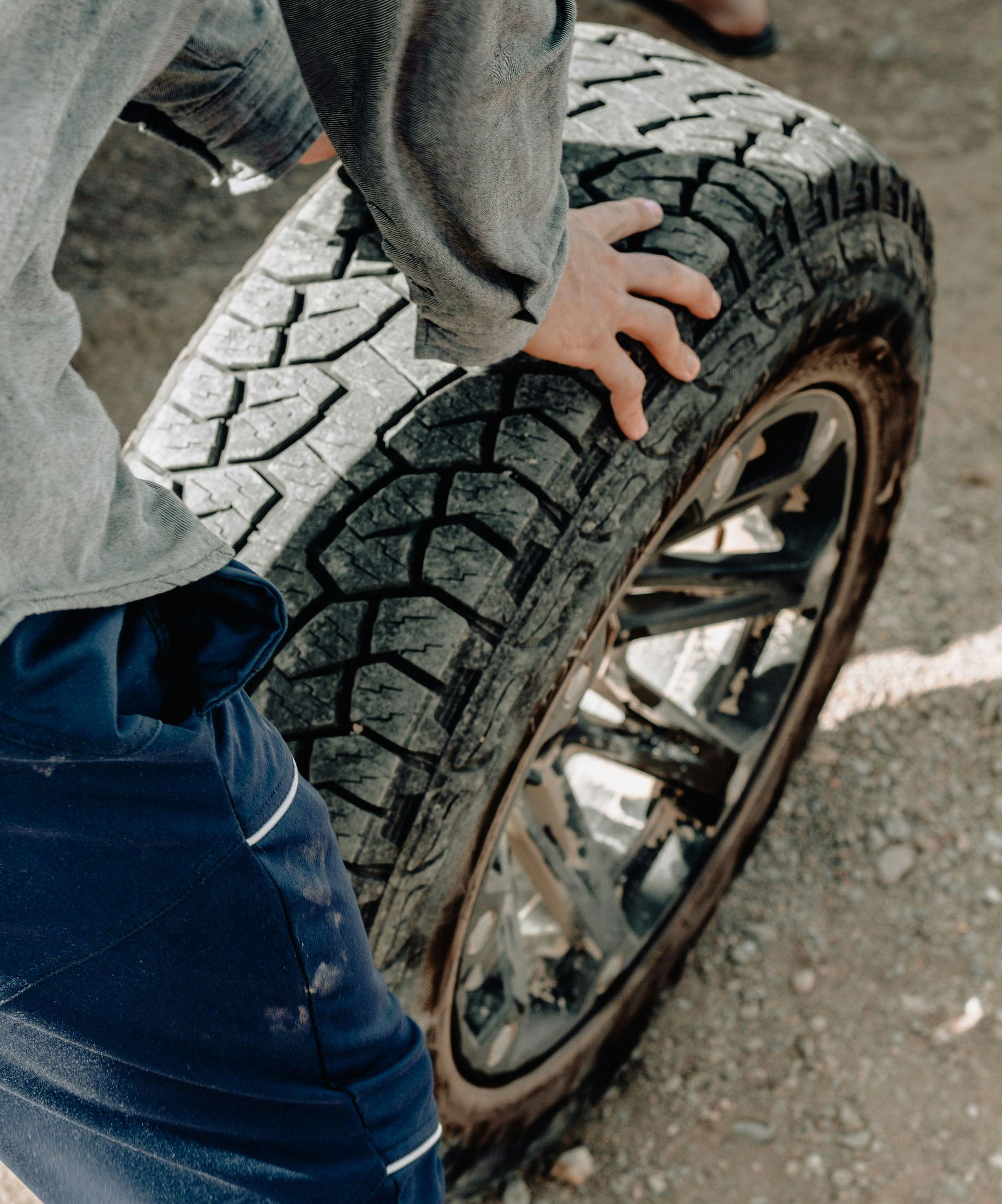 A person wearing a grey shirt and blue pants grips a large, treaded tire on a metal rim on a dirt surface.