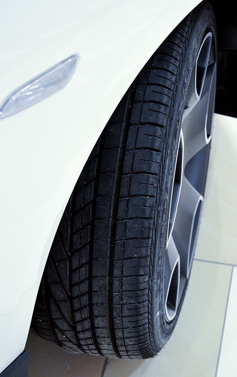 White car's front tire and alloy wheel close-up. The black tire has tread, and the wheel has a silver rim.