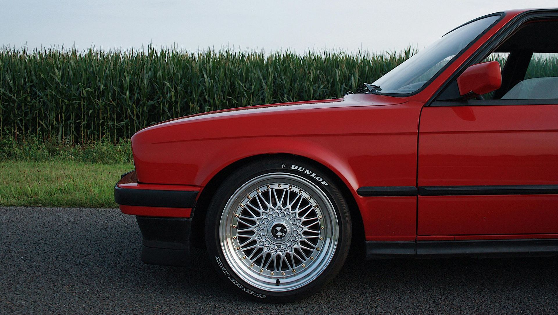 A red car is parked in front of a corn field
