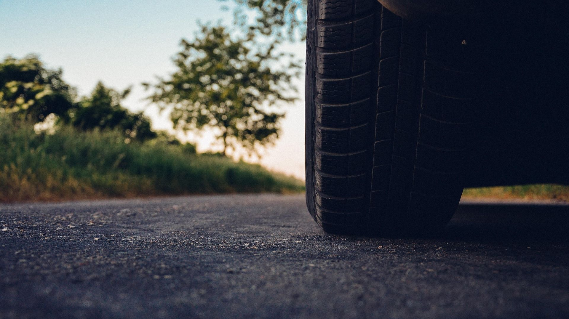 Close-up of a car tire on an asphalt road; blurred background shows trees and a partly cloudy sky.