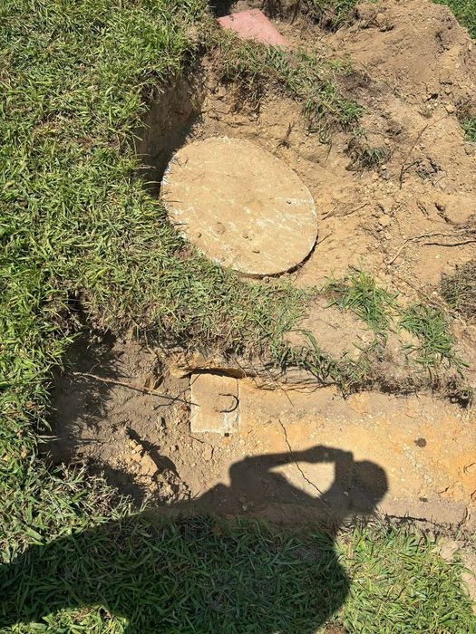 Round concrete lid and rectangular block in dirt and grass, near a shadow.