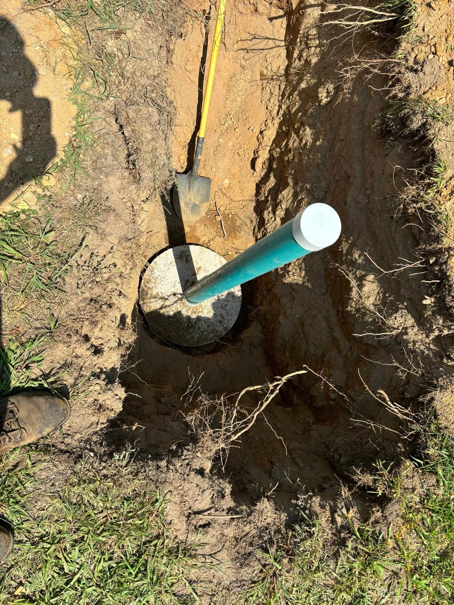A well with a green pipe and a white cap, surrounded by dirt and grass; a shovel is nearby.