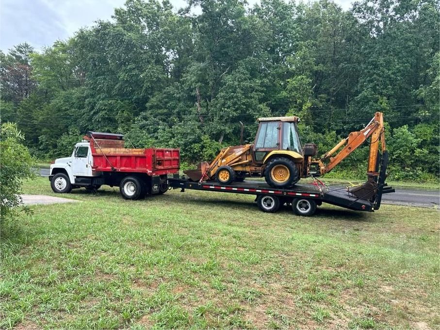 Truck towing a trailer with a yellow backhoe on it in a grassy area, trees in the background.