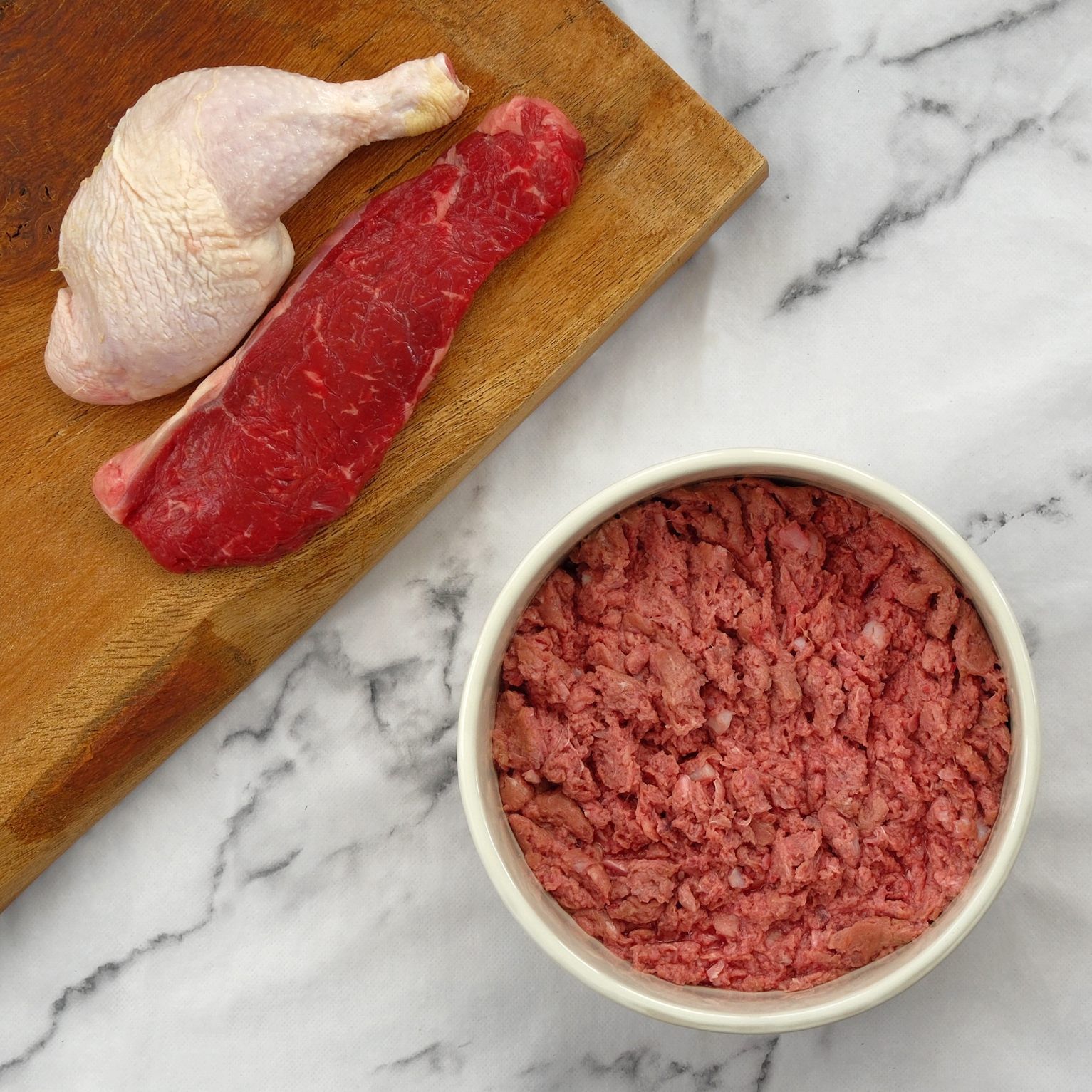 Raw chicken leg, beef strip, and a bowl of ground meat on a wooden board.