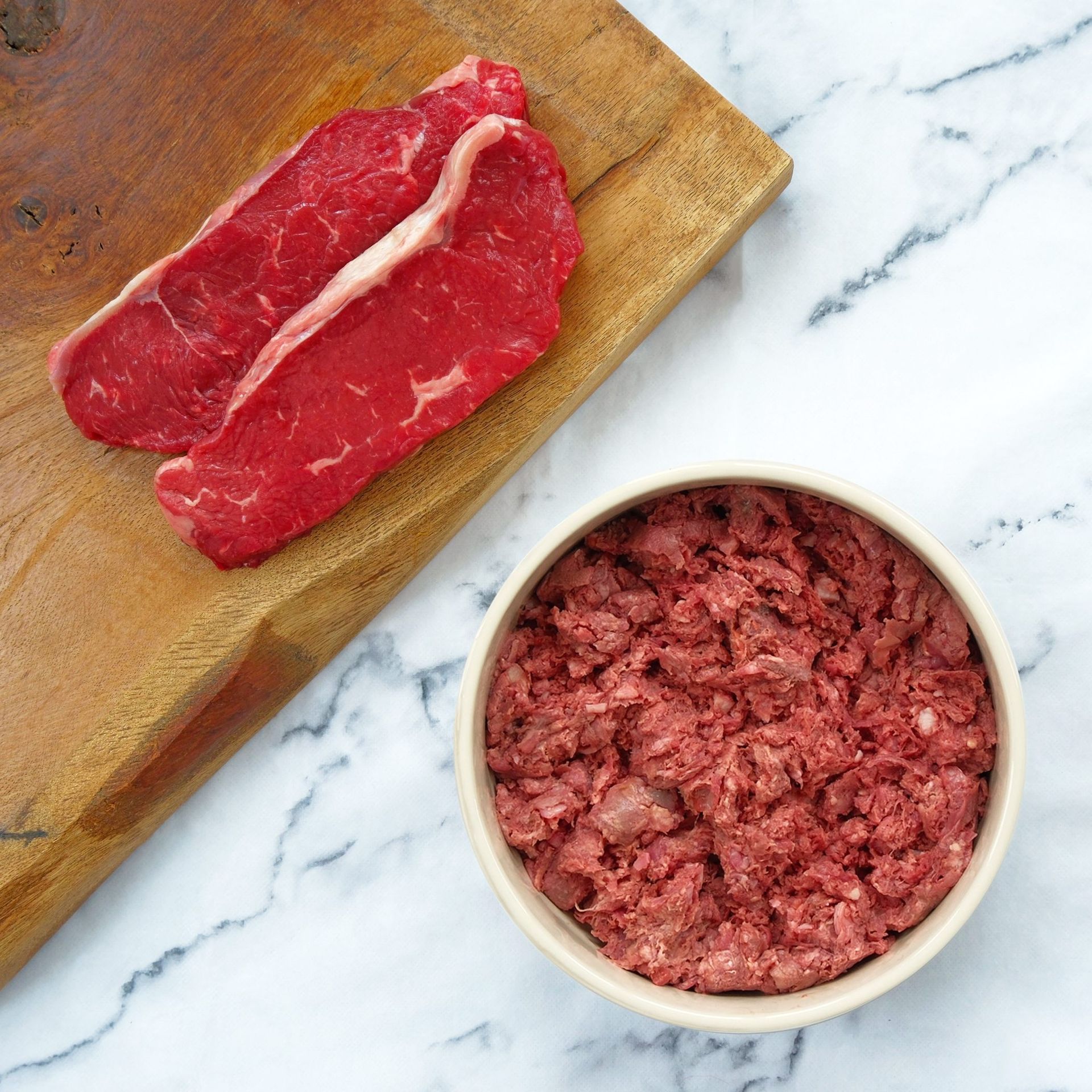 Raw red meat, two steaks on a cutting board, and ground meat in a bowl, on a marble surface.