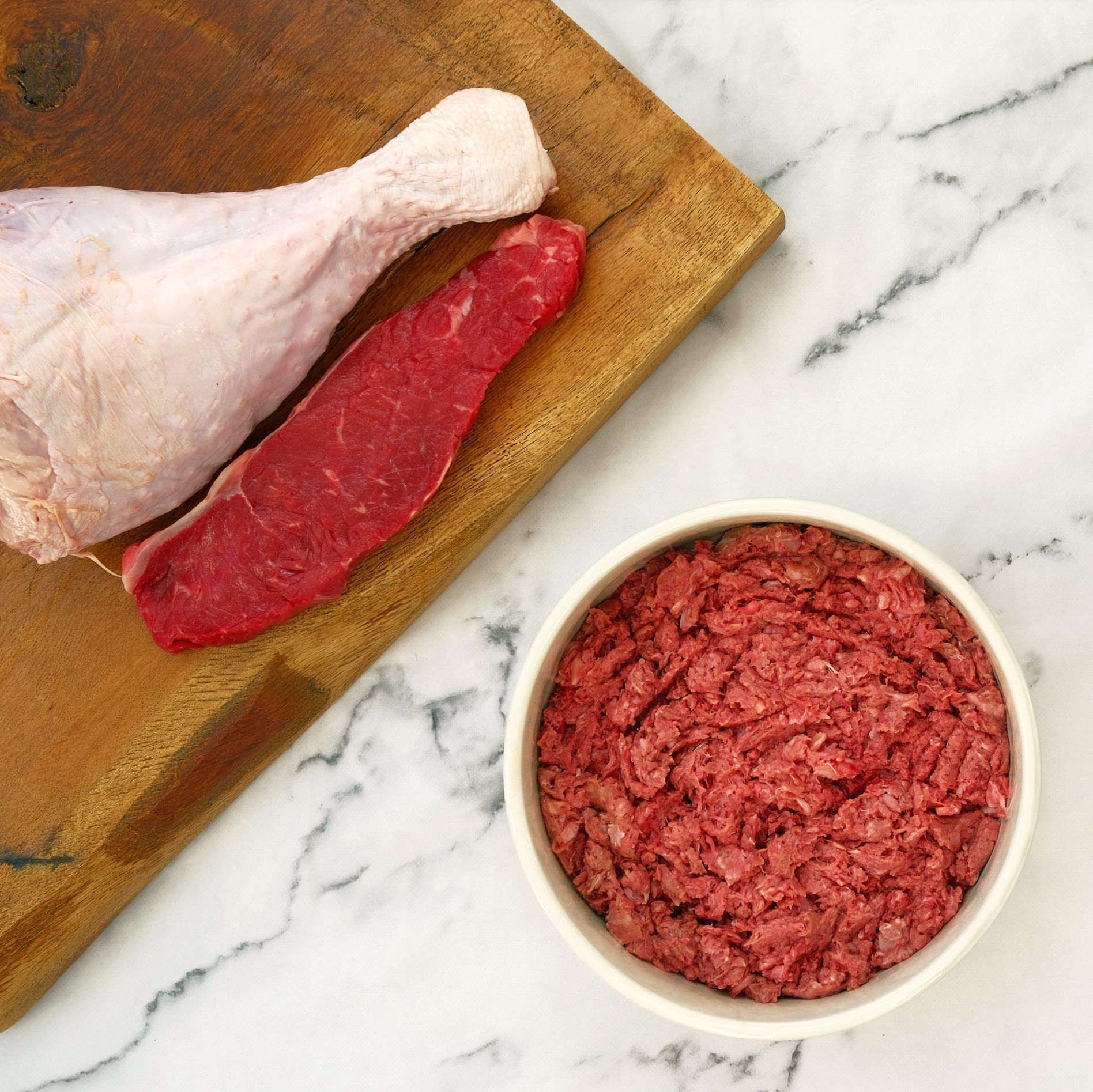 Raw meat ingredients: a turkey leg, beef steak, and ground meat in a bowl on a cutting board.
