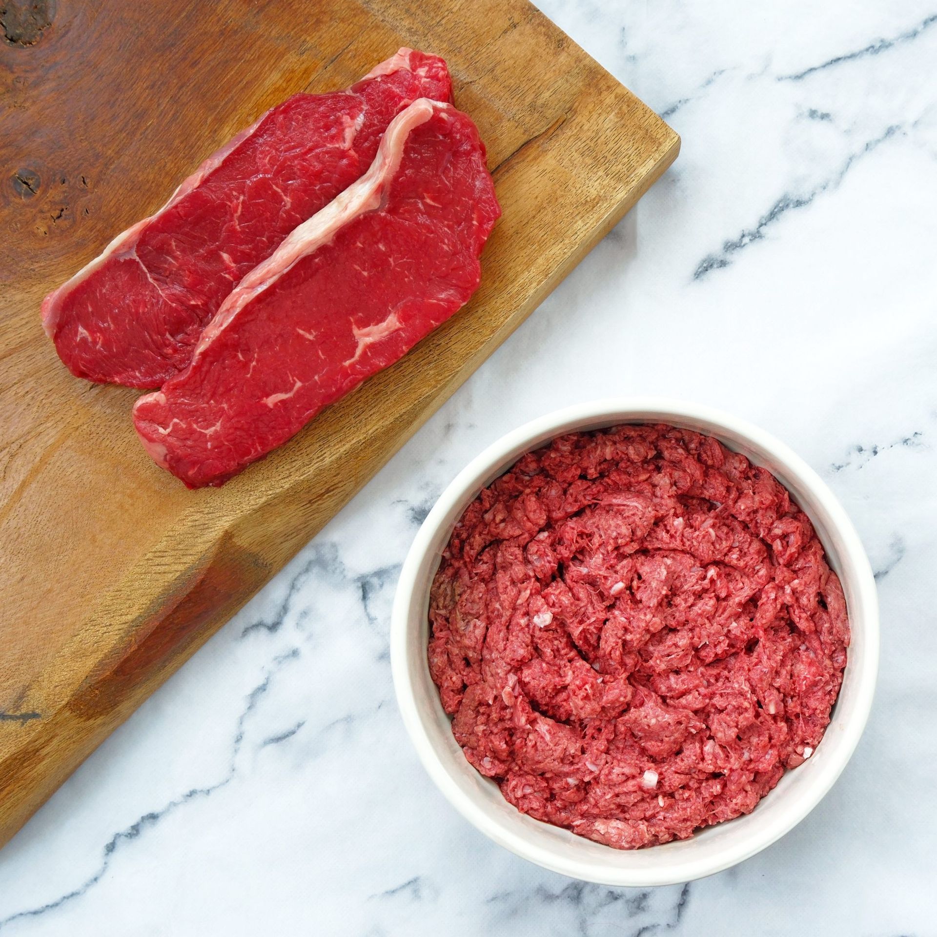 Two raw steaks and a bowl of ground meat on a wooden cutting board and marble surface.