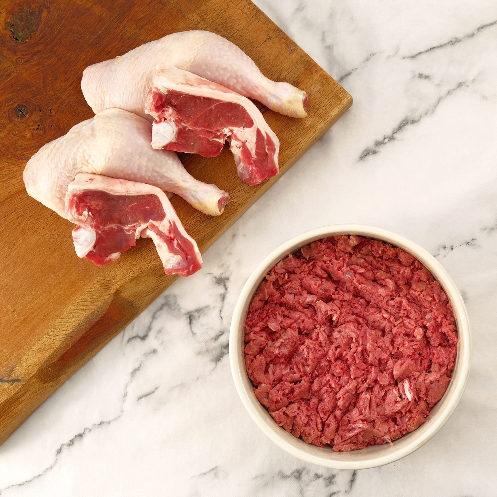 Raw lamb leg quarters and ground lamb in a bowl, on a wooden cutting board and marble surface.