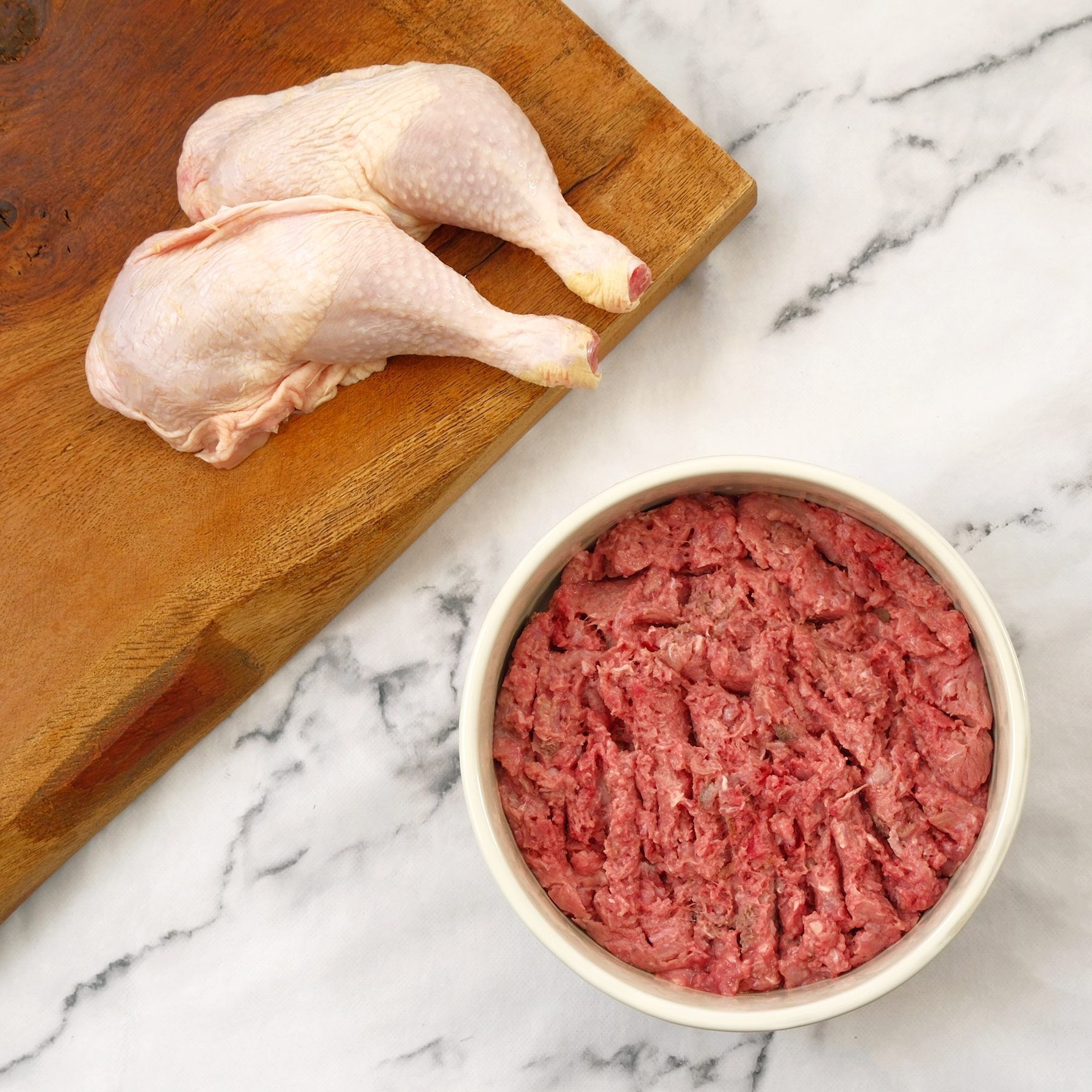 Raw chicken legs on a wooden board, next to a bowl of ground raw meat on marble.