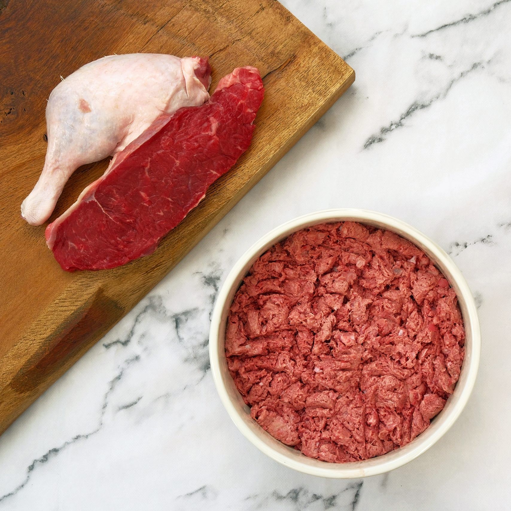 Raw meat ingredients: duck leg, steak, and ground meat in a bowl on a wooden board.