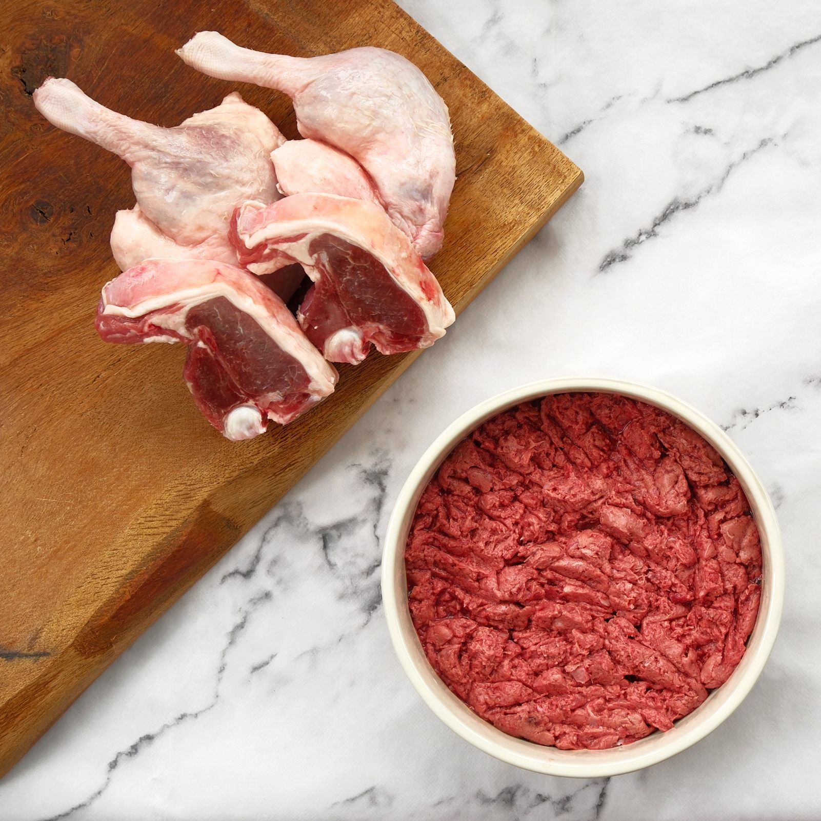 Duck legs and ground meat in a bowl on a wooden board against a marble surface.