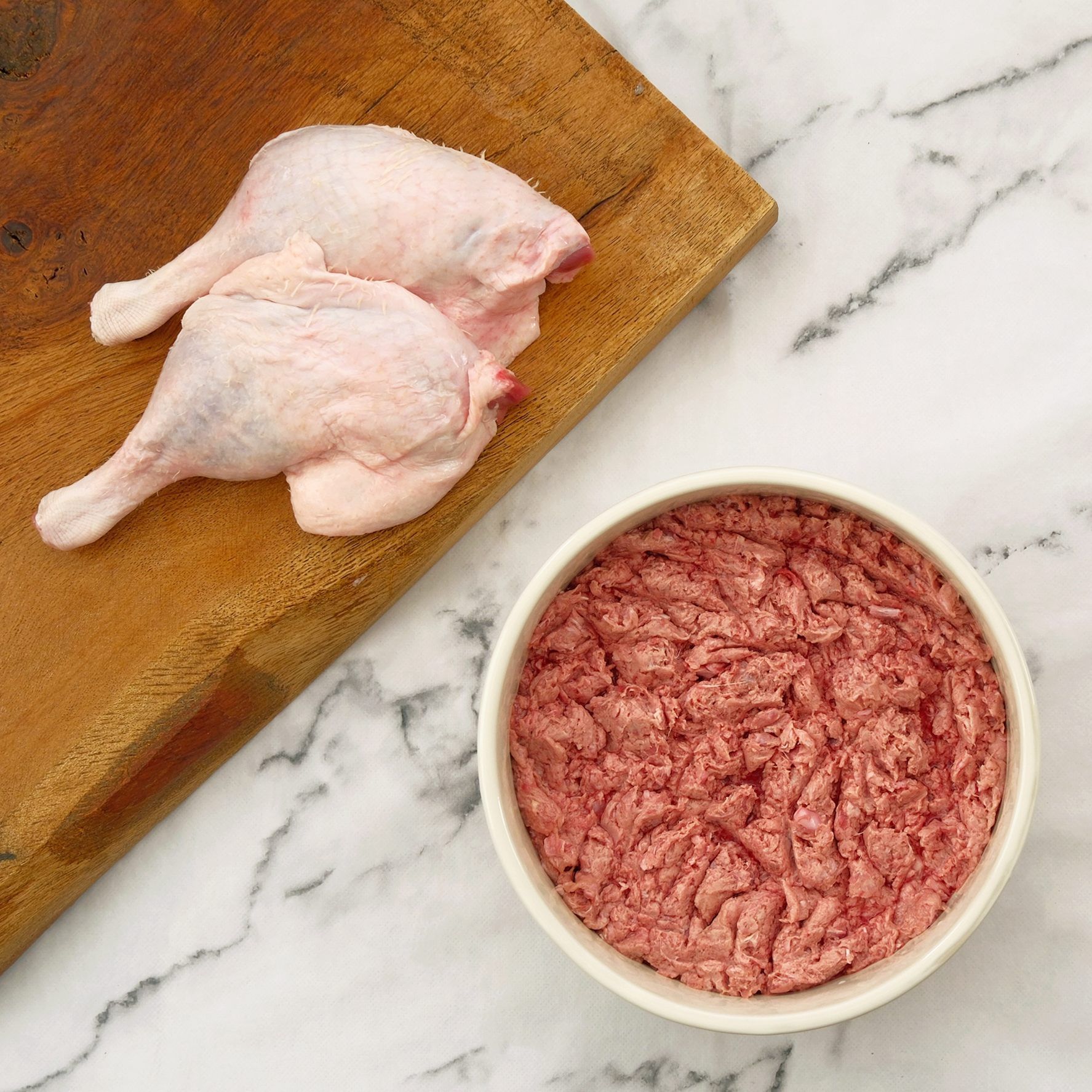 Duck legs on a wooden board next to a bowl of ground duck meat.