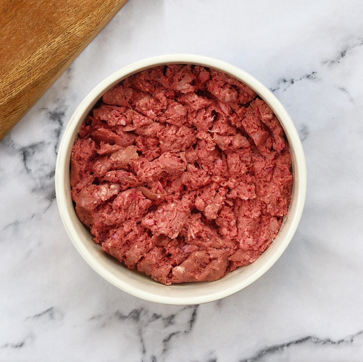 Bowl of raw, ground meat on a marble surface. Brown cutting board in the corner.