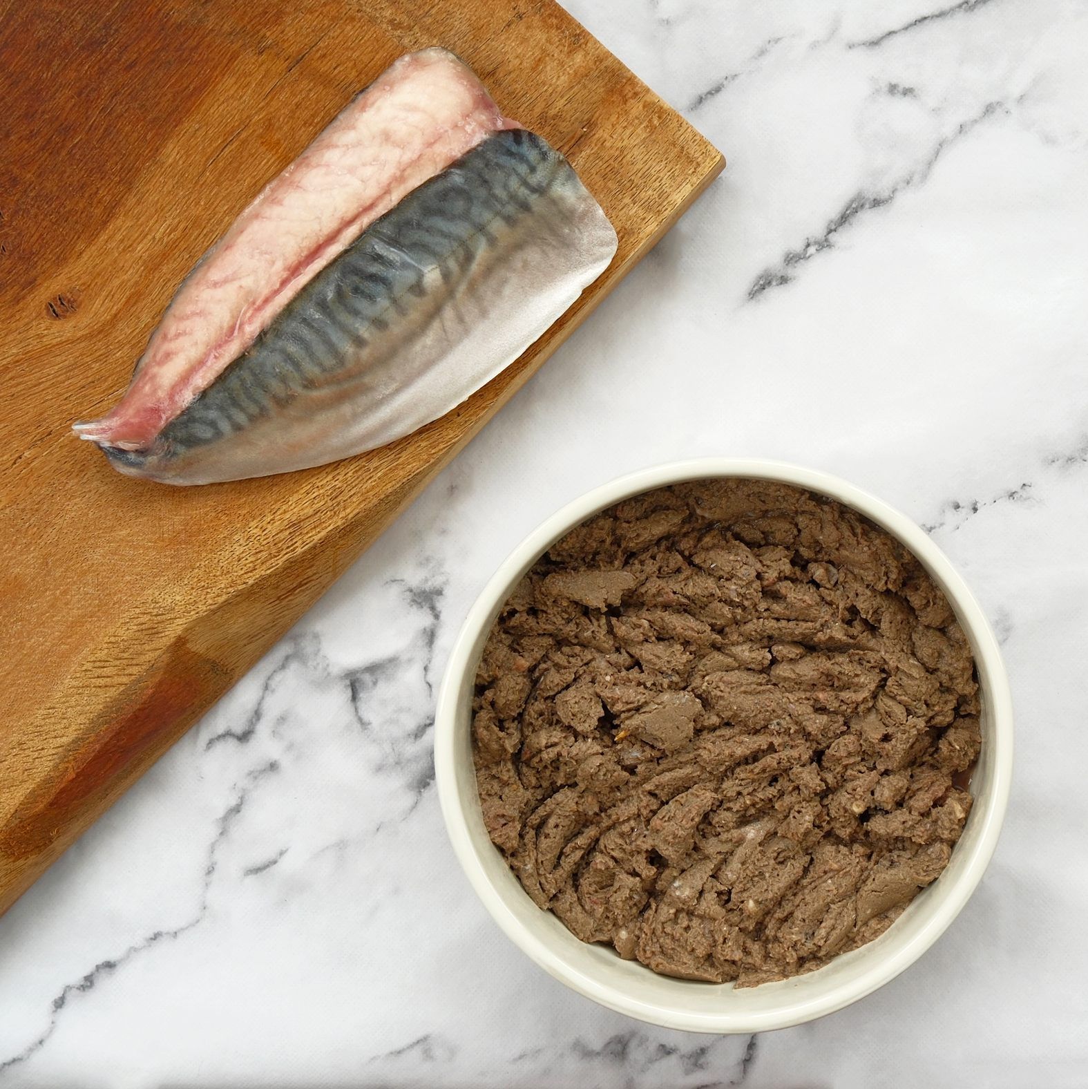 Raw fish fillet on wooden board next to a bowl of brown wet cat food, on a marble surface.