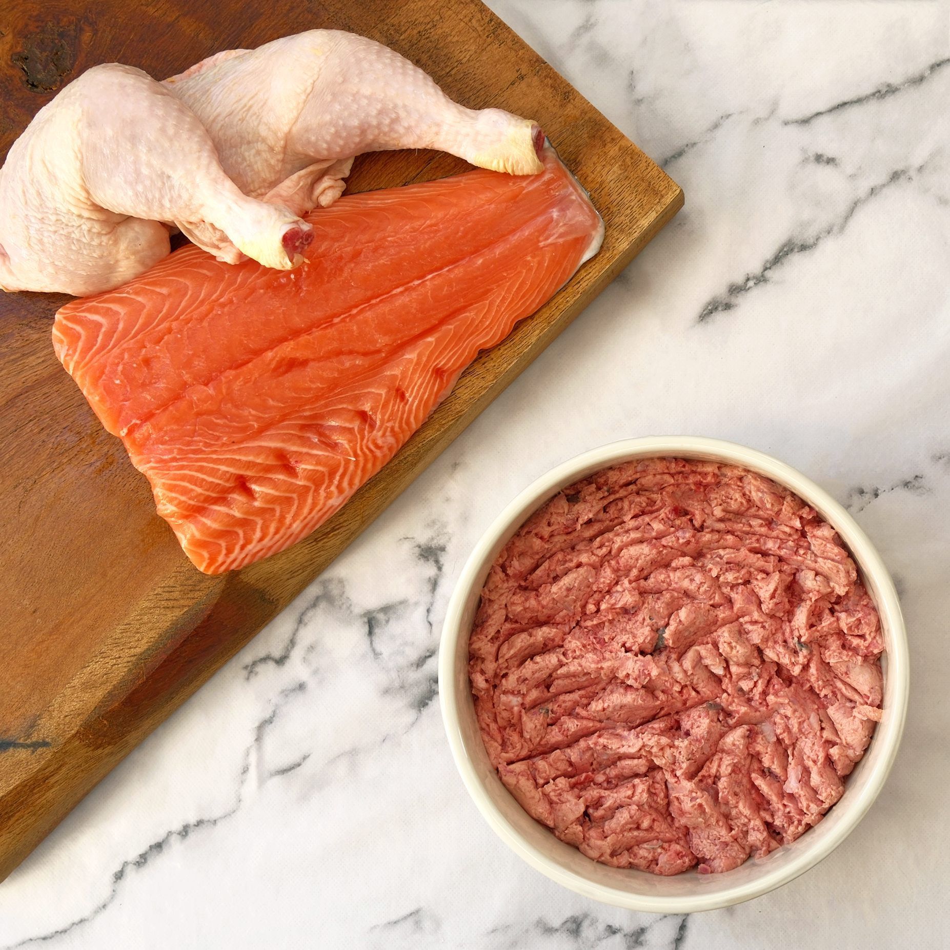 Raw chicken leg quarter, salmon fillet, and ground meat in a bowl on a wooden board.