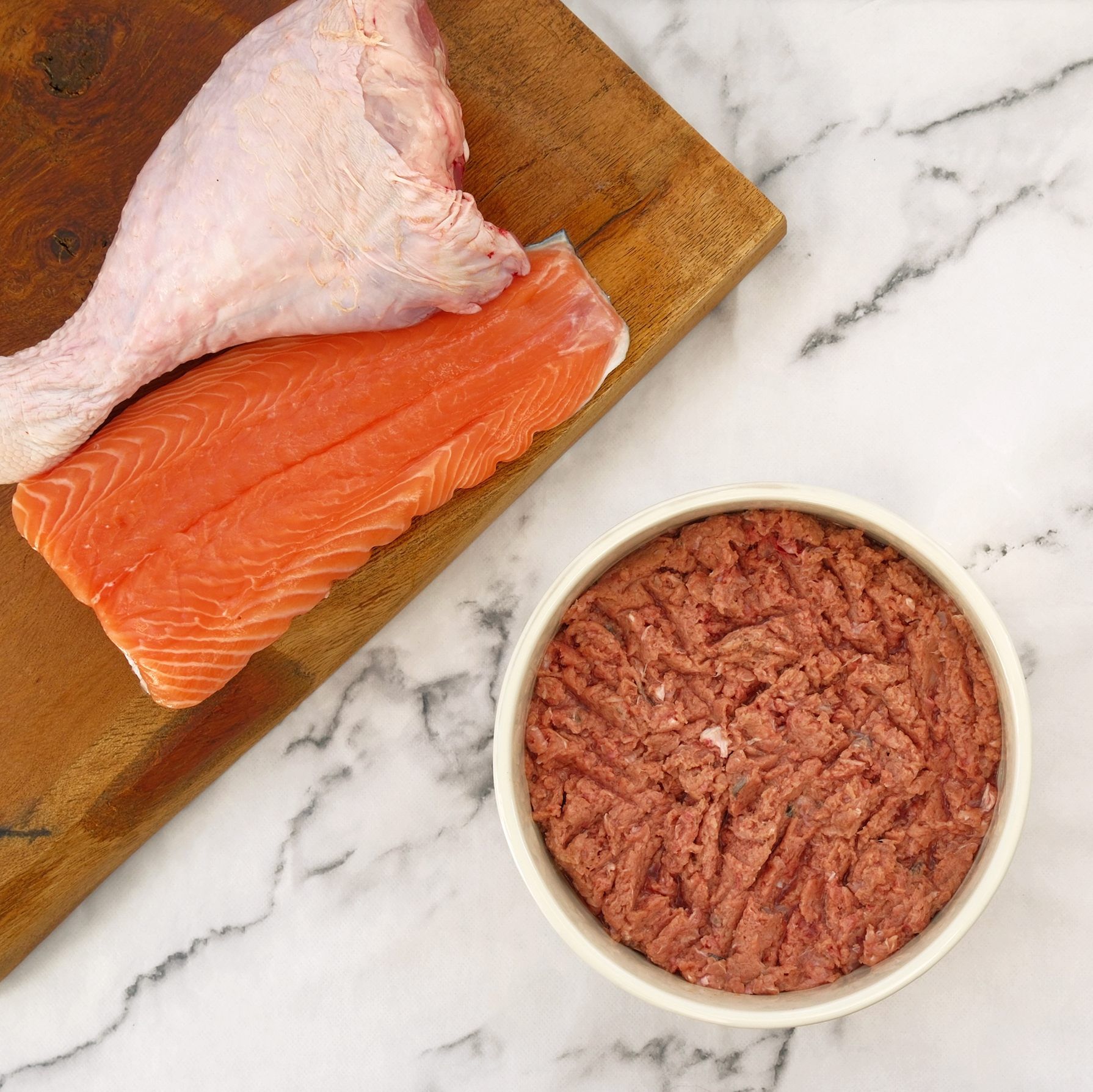 Raw turkey leg, salmon fillet, and ground meat in a bowl on a wooden board.