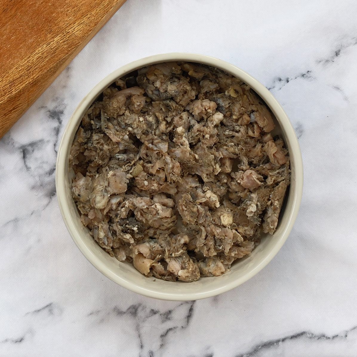 Bowl of chunky, gray-brown pet food on a marble surface, with a wooden item partially visible.